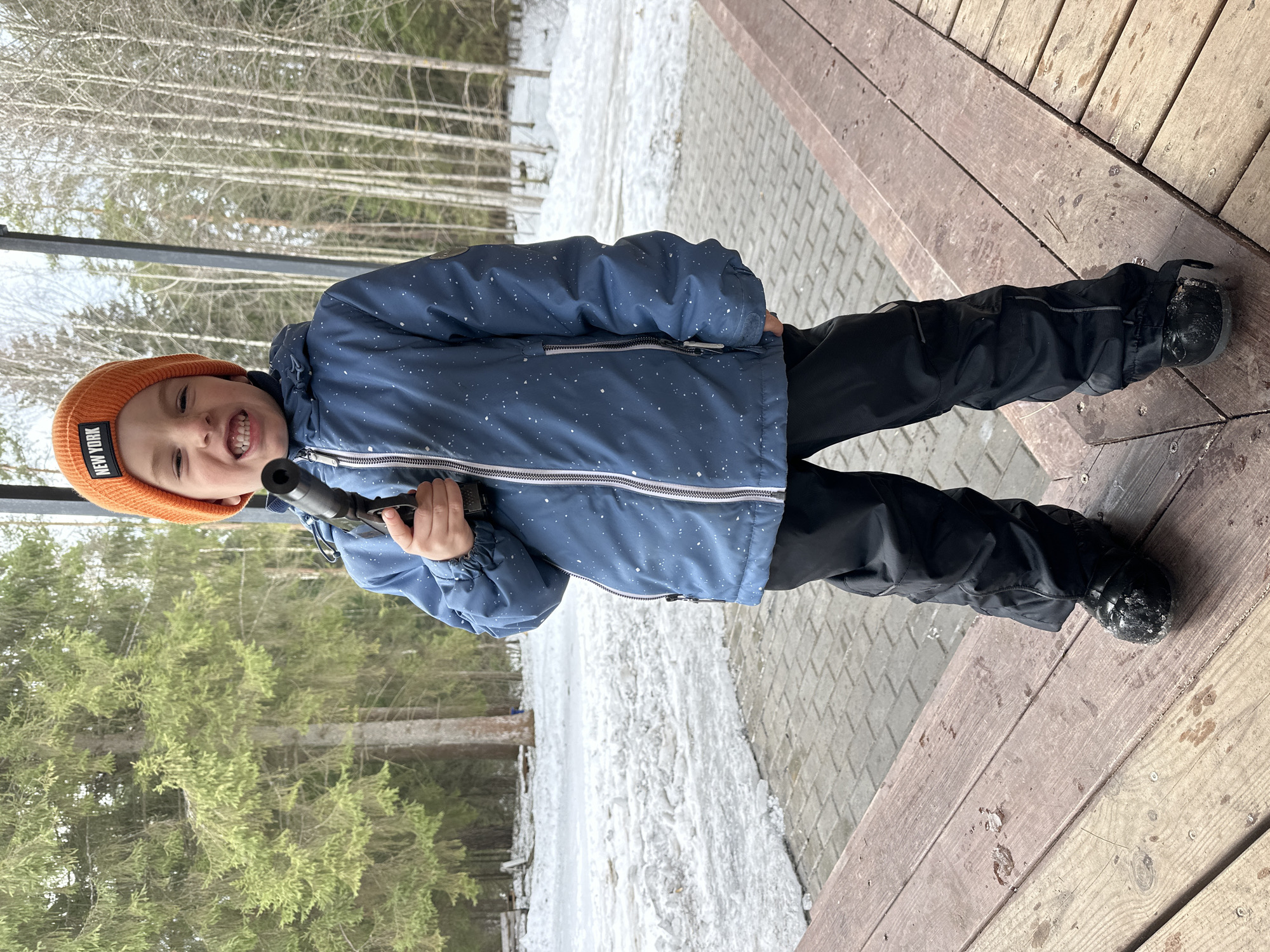 A young child stands outdoors, holding a dark object resembling a toy gun. He wears a blue winter jacket with white speckles, black pants, and black boots with some snow on them. He also wears an orange beanie with the words "NEW YORK" on it. The child is on a wooden deck, with snow and ice visible in the background, along with trees. The location is in Прикамский, Russia.