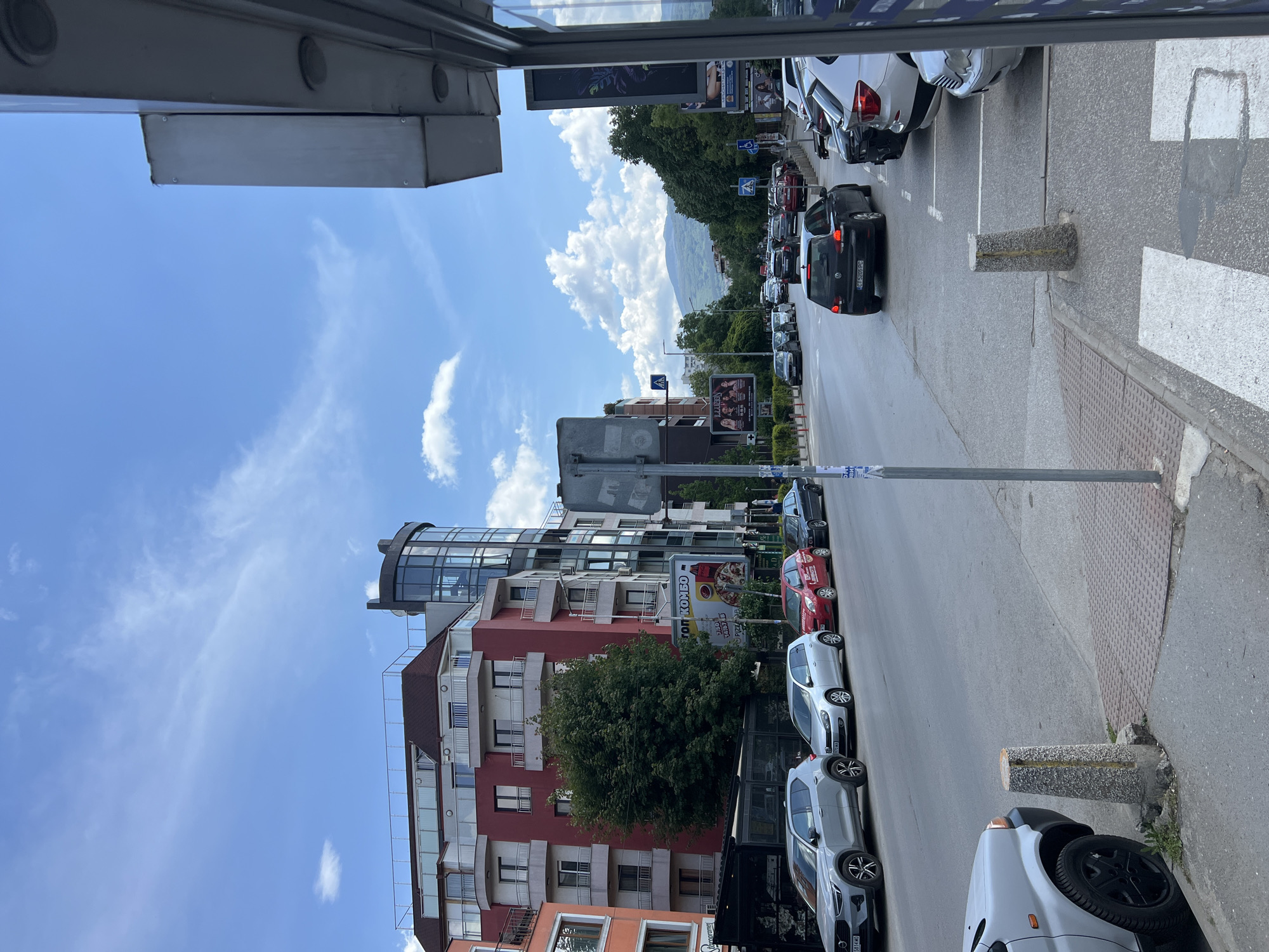 The image depicts a street scene in Sofia, Bulgaria, with buildings on the left and cars parked on the right. The sky is blue with scattered clouds. A mountain is visible in the distance. Buildings vary in height and color, including red and orange. A structure with a rounded, glass-enclosed top is among the buildings. Several billboards are placed along the street. Cars are parked along the right-hand side of the street. Parking is marked by concrete bollards. A pedestrian crossing is present on the right. The photo is taken from within a bus or similar vehicle.