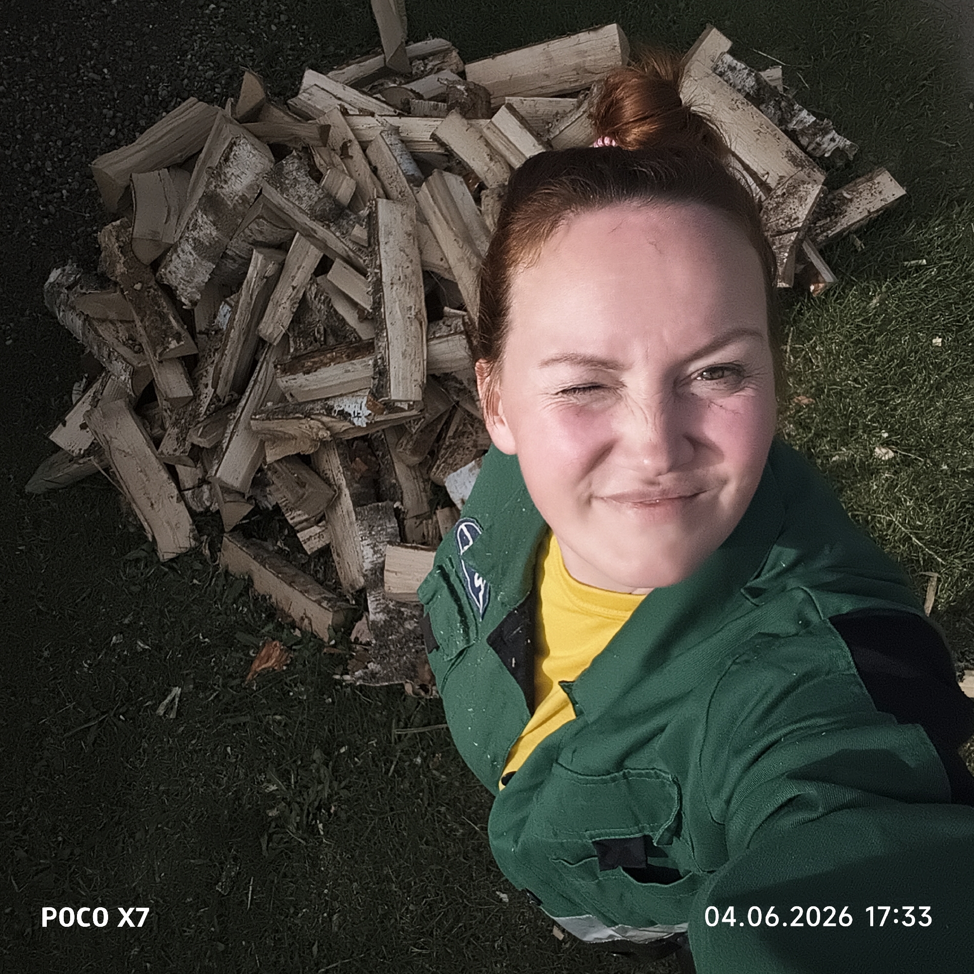 A young woman is captured in a high-angle selfie outdoors in Chaikovsky, Russia, on June 4, 2026, at 17:33. She is looking at the camera, winking with her right eye and smiling playfully. Her light reddish-brown hair is pulled back into a bun. She wears a bright yellow t-shirt underneath a dark green work jacket or overall, which has black sections and a white emblem visible on the left sleeve. Small specks of white material, possibly sawdust or paint, are visible on her jacket.
Behind and to her left, a substantial pile of chopped firewood is scattered across a grassy area. The logs are of various sizes, with some retaining their light-colored, peeling bark, characteristic of birch. The ground is covered in green grass, suggesting an outdoor, possibly rural or semi-rural setting. The scene is illuminated by bright sunlight, indicating a clear, late afternoon or early evening sky, which casts shadows and highlights on the woman's face.
Visible text on the image includes "POCO X7" in the bottom left corner, indicating the device used for the photograph, and the date and time "04.06.2026 17:33" in the bottom right corner. The overall mood conveyed by the woman's expression is cheerful and content.