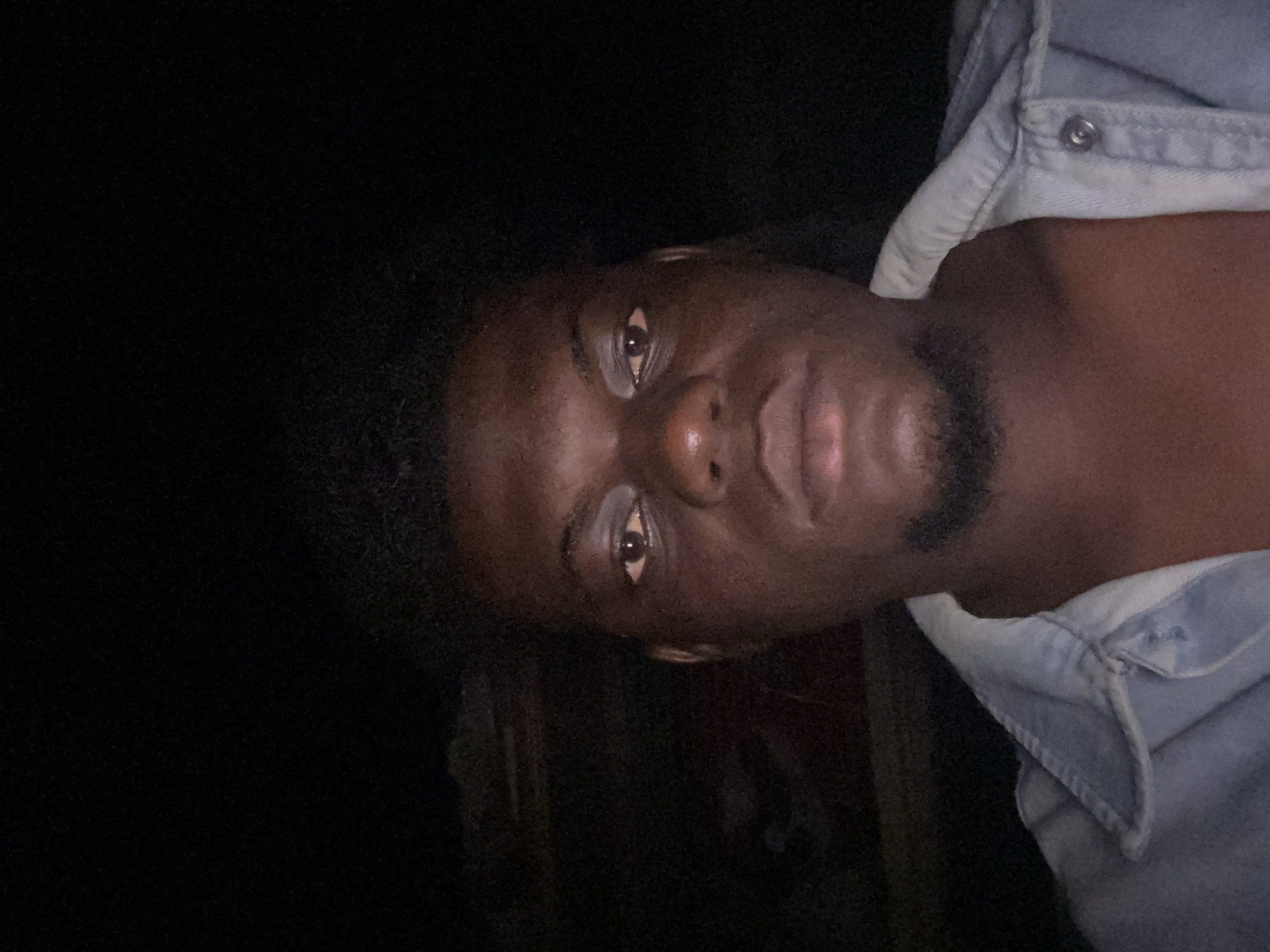 The image presents a close-up portrait of a dark-skinned individual, oriented sideways (rotated 90 degrees clockwise from an upright position). The person, likely male, is looking directly at the camera with a neutral to slightly serious expression.
The subject's face occupies the right half of the frame, illuminated by a direct light source, possibly a flash, which creates strong highlights on the forehead, bridge of the nose, and lips. The eyes are dark with visible whites and distinct catchlights, reflecting the light source. Dark, curly hair is visible on the left side of his head, and he sports a short, dark goatee or beard around his chin and mouth area.
He is wearing a light-colored, collared shirt, which appears to be made of denim or chambray fabric. A silver-toned snap button is visible near the collar on what would be his right shoulder in an upright view.
The background is predominantly dark, almost completely black, indicating a very low-light environment, possibly indoors or at night. There are no discernible details or objects in the background due to the extreme darkness.
No visible text is present in the image. The overall impression is a direct, intimate portrait captured in challenging lighting conditions.