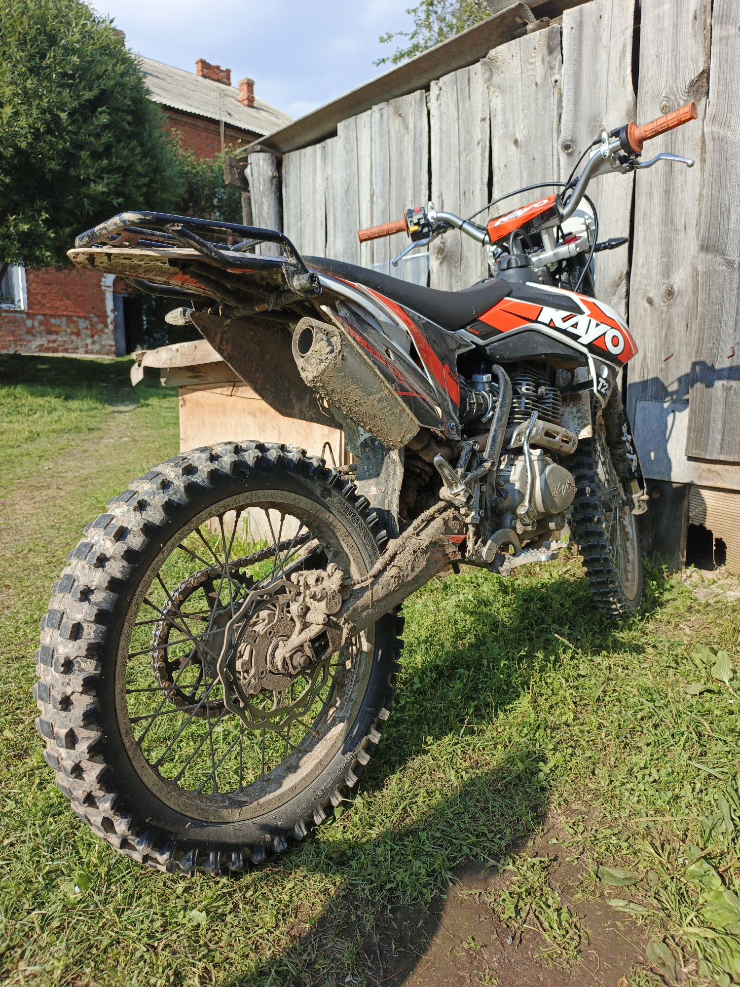 A dirt bike, branded with "KAYO" in white and red lettering, is parked on a grassy area next to a weathered wooden fence. The bike is heavily coated in mud, suggesting recent off-road use. It features black and orange body panels, knobby tires, and an exposed engine. In the background, a red brick building with a grey roof and chimneys is visible, along with a large green tree. The scene is set outdoors in what appears to be a rural or semi-rural environment in Medvedyovka, Russia. The lighting indicates it is daytime. There are no people present in the image.