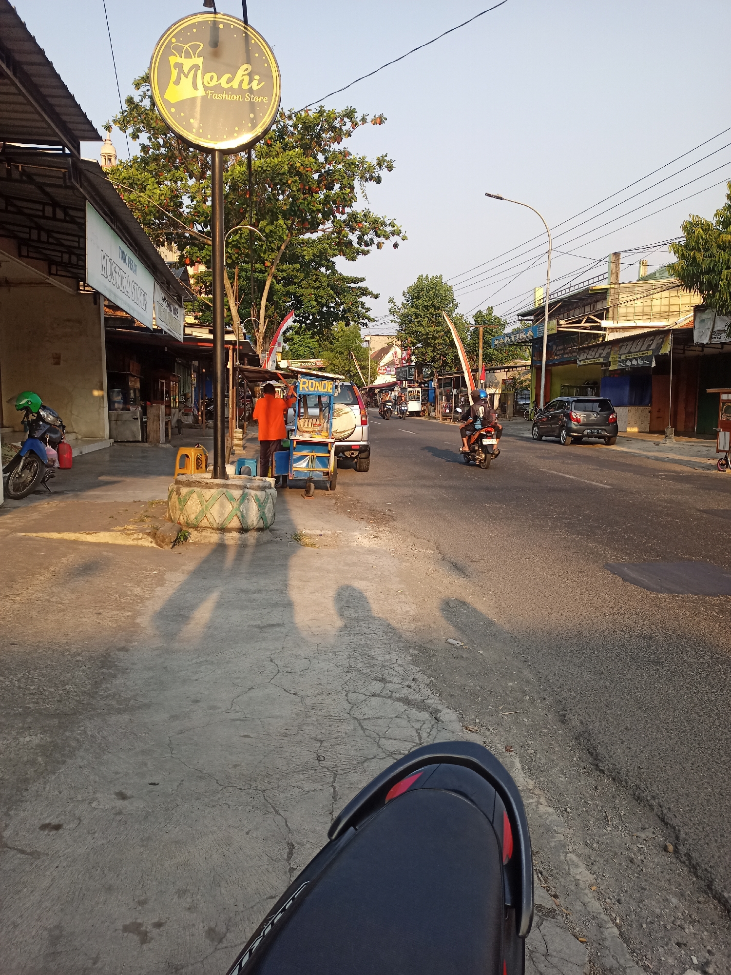 The image captures a street scene in Gemolong, Indonesia, at what appears to be late afternoon given the long shadows. The viewpoint is from the seat of a motorcycle, looking forward. On the left side of the street, a blue motorcycle with a green helmet is parked in front of a building with an awning. A "Mochi Fashion Store" sign is prominently displayed on a lamppost. Further down the sidewalk, a person in an orange shirt is attending to a mobile food cart. Several small yellow and blue stools are arranged around the cart. A white car with a blue and yellow cart is parked to the right of the food vendor. The street itself is paved and shows some signs of wear. Vehicles, including another motorcycle with two occupants and a silver car, are driving on the road. Buildings line the right side of the street, with various awnings and signs indicating businesses. Trees with green foliage are visible throughout the scene, some with red fruit. The sky is clear and light blue, suggesting a sunny day.