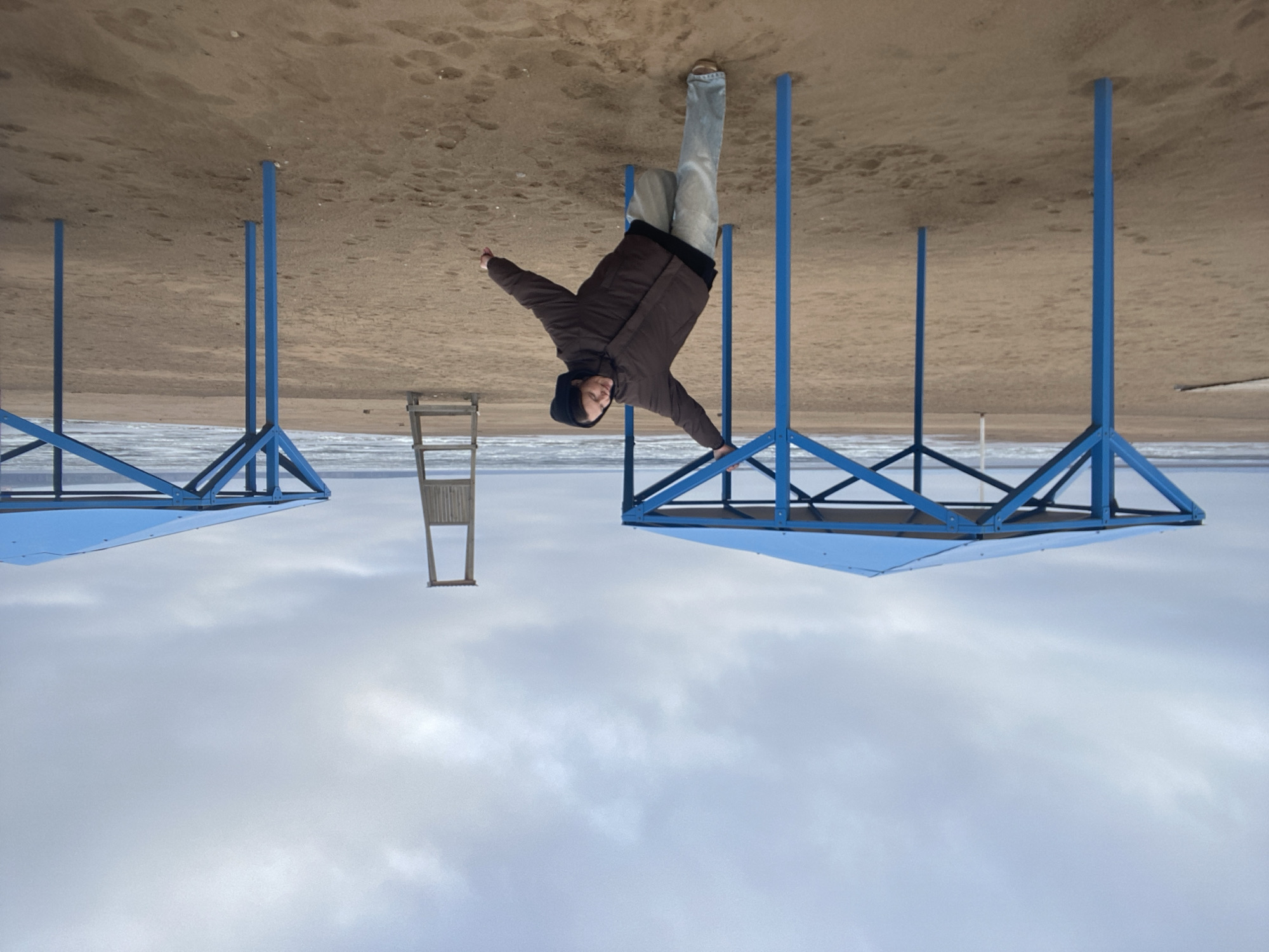 This inverted image displays a surreal scene on what appears to be a beach. In the center, a person is depicted upside down, seemingly standing on a cloudy sky. They are wearing a brown jacket with a dark hood and light-colored pants. Their arms are outstretched, with one hand resting on a blue metal structure. The person's expression is neutral to slightly smiling.
Above the person, where the sky would normally be, is a sandy ground with visible footprints and some small debris. Below the person, acting as the ground in this inverted view, is a vast expanse of light grey, cloud-filled sky. Several blue metal structures, resembling frames for beach umbrellas or gazebos, are prominently featured, also appearing upside down. One is near the central figure, another is partially visible on the far left, and parts of others are seen to the right. A wooden chair is also inverted in the middle-left background. A narrow band of water, likely a sea, lake, or large river, is visible horizontally between the sandy 'sky' and the cloudy 'ground'.
When viewed in its intended orientation, the image captures a person striking a playful or artistic pose on a sandy beach next to a body of water, under a cloudy daytime sky. The blue structures are beach equipment, and the wooden chair is a typical beach accessory. There is no visible text in the image. The setting is a beach environment, consistent with Chaikovsky, Russia, implying a large body of water nearby.