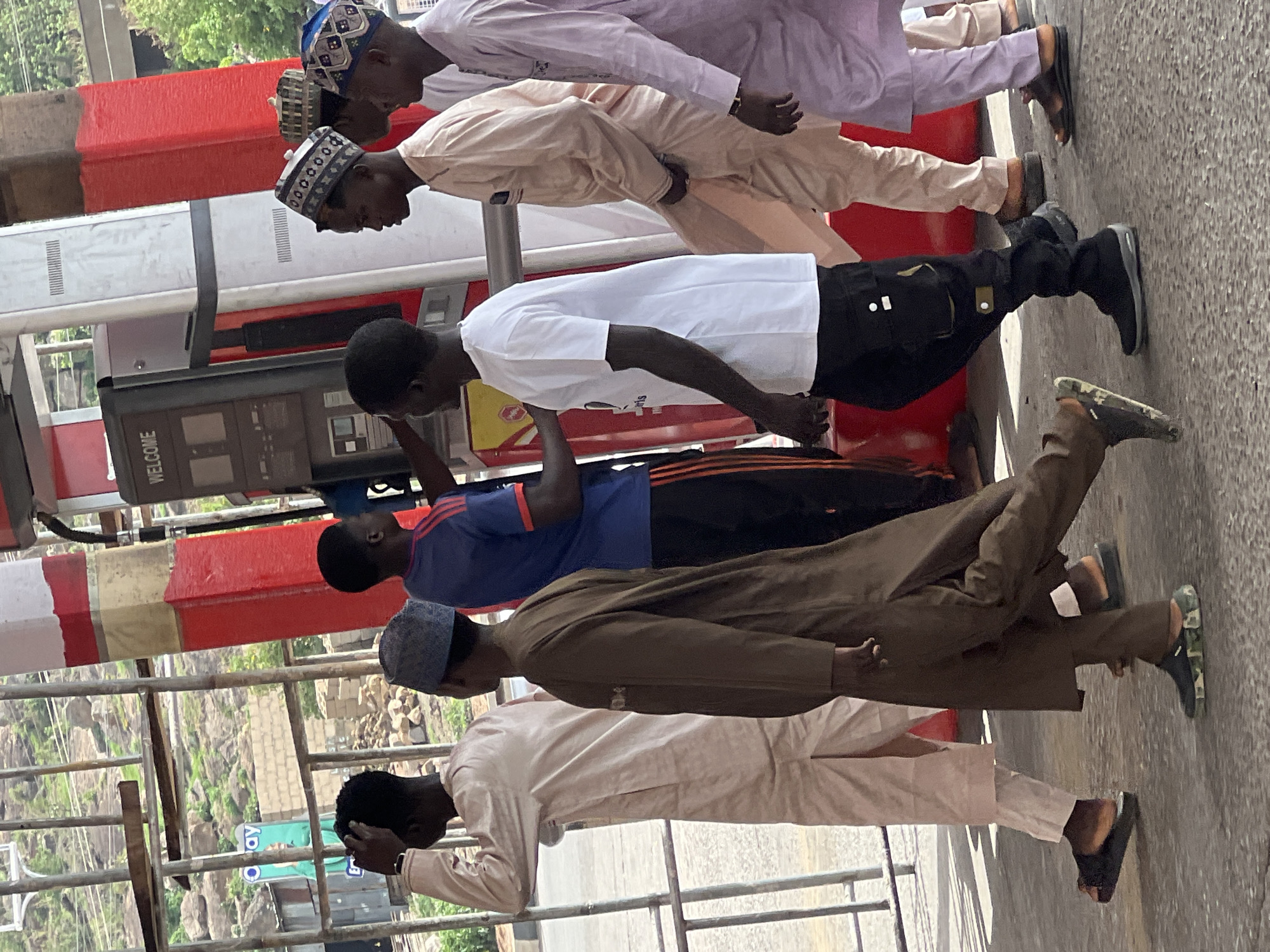 A medium shot captures six men standing in a line on what appears to be a paved or asphalt surface in Gura Top, Nigeria. The men face right, and their heads are bowed, creating a sense of solemnity or reverence.
Clothing varies among the men. The first man wears a light purple robe and a patterned hat. The second man is dressed in a light tan robe with a similar hat. The third man has on a white t-shirt and black pants. The fourth man is wearing a blue shirt with orange stripes on the sleeves, along with black pants. The fifth man is in a brown robe and a blue hat. The sixth man has on a light tan robe.
Behind the men, a red and white structure is visible, possibly part of a building or infrastructure, and a fuel pump with the word "WELCOME" on its display. A metal railing runs parallel to the men. In the background, a hillside with vegetation and a few buildings can be seen. The lighting suggests a sunny day.