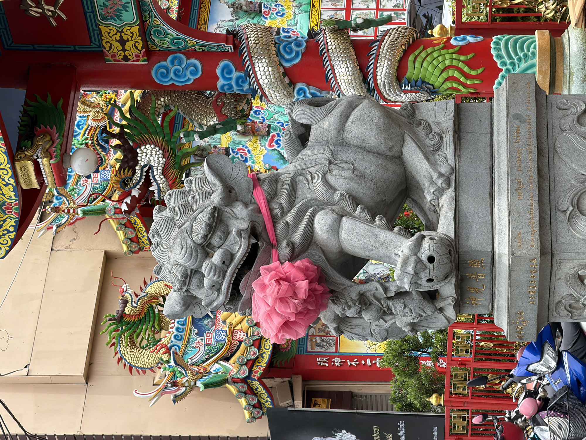 A stone statue of a foo dog or guardian lion is positioned in the foreground, adorned with a pink fluffy ornament and a pink ribbon. Behind it, a vibrant red gateway with ornate dragon and cloud carvings is visible. The gateway leads into a courtyard with a building featuring intricate, colorful architectural details, including dragons and traditional motifs. The overall scene suggests a Chinese temple or shrine in a bustling urban environment, likely in Chiang Mai City Municipality, Thailand. In the lower left corner, parts of motorcycles are visible, indicating their proximity to the street.