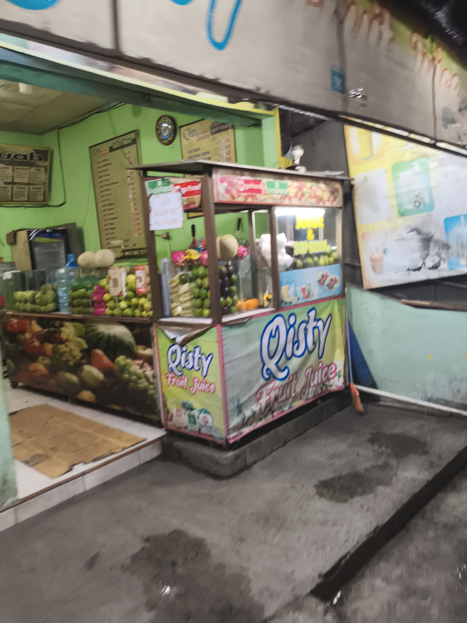 This appears to be a fruit juice stand, located in Depok, Indonesia, based on signage. The stand is situated along a street or sidewalk. A light green building houses the main part of the operation. Fruits and supplies are displayed behind the counter. The front of the stand has a sign that says "Qisty Fruit Juice". The area outside the stand is paved and there's some dampness on the ground. The photo is taken at night or in a dimly lit area.