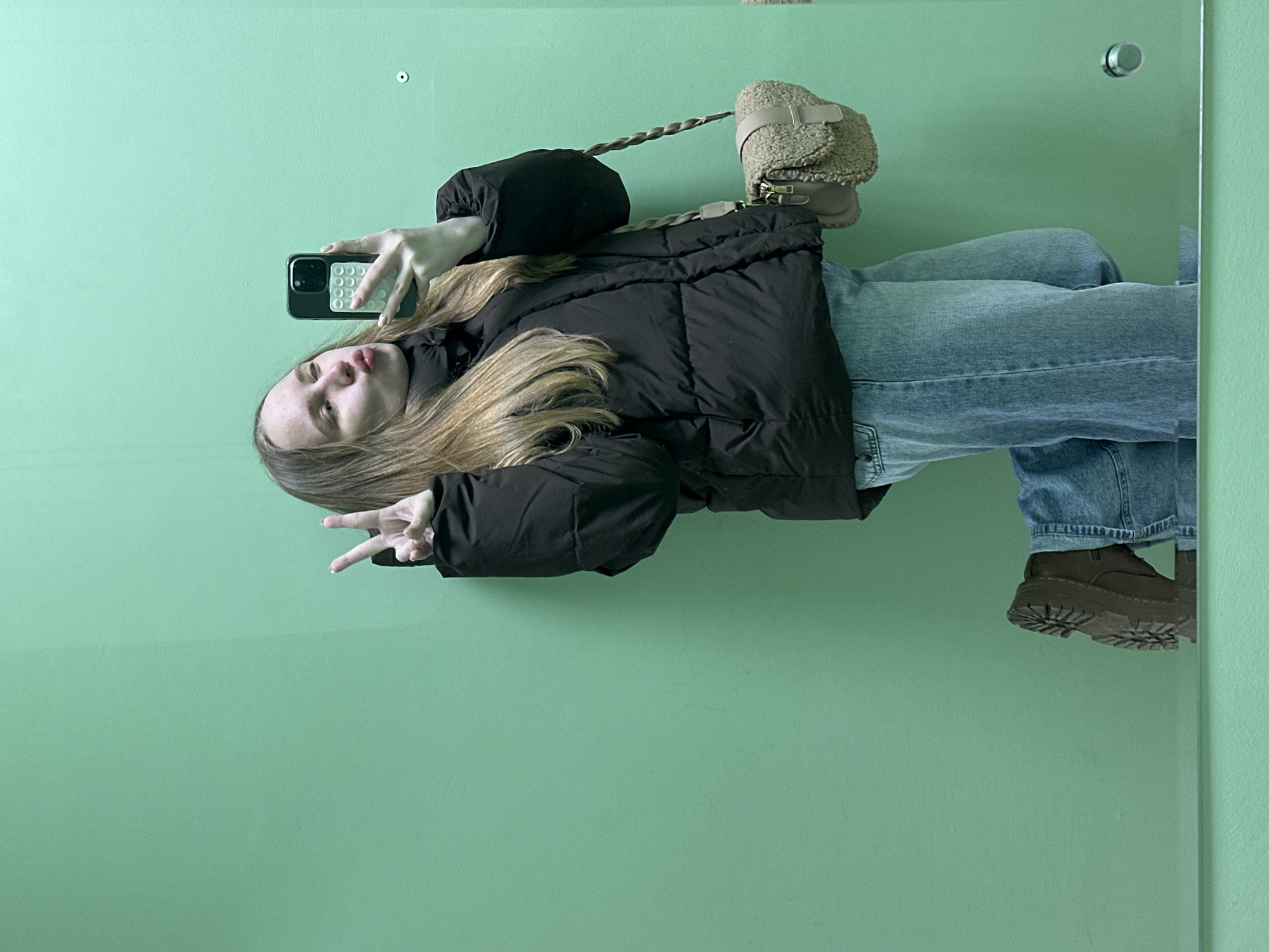 A young person, appearing to be female, is captured in a mirror selfie against a plain, light green wall. They have long, light brown hair flowing over their shoulders and are dressed in a black puffer jacket, light blue denim jeans, and chunky dark brown boots with thick, ridged soles.
The individual is holding a black smartphone, featuring a distinctive white, textured case, up with their right hand to take the photo. Their left hand is extended to the side, forming a peace sign. Their facial expression is a subtle pout, as they look towards the phone screen. A light beige or cream-colored fuzzy shoulder bag with a braided strap is visible, hanging from their right shoulder.
The scene is indoors, reflected in a large mirror, the edge of which is visible on the right side. The background behind the mirror is a solid, clean light green wall. Small, circular fixtures are present on the wall: a white one on the left and a metallic one on the right, possibly functioning as mirror mounts or decorative elements. The overall atmosphere is casual and contemporary. There is no visible text in the image.
