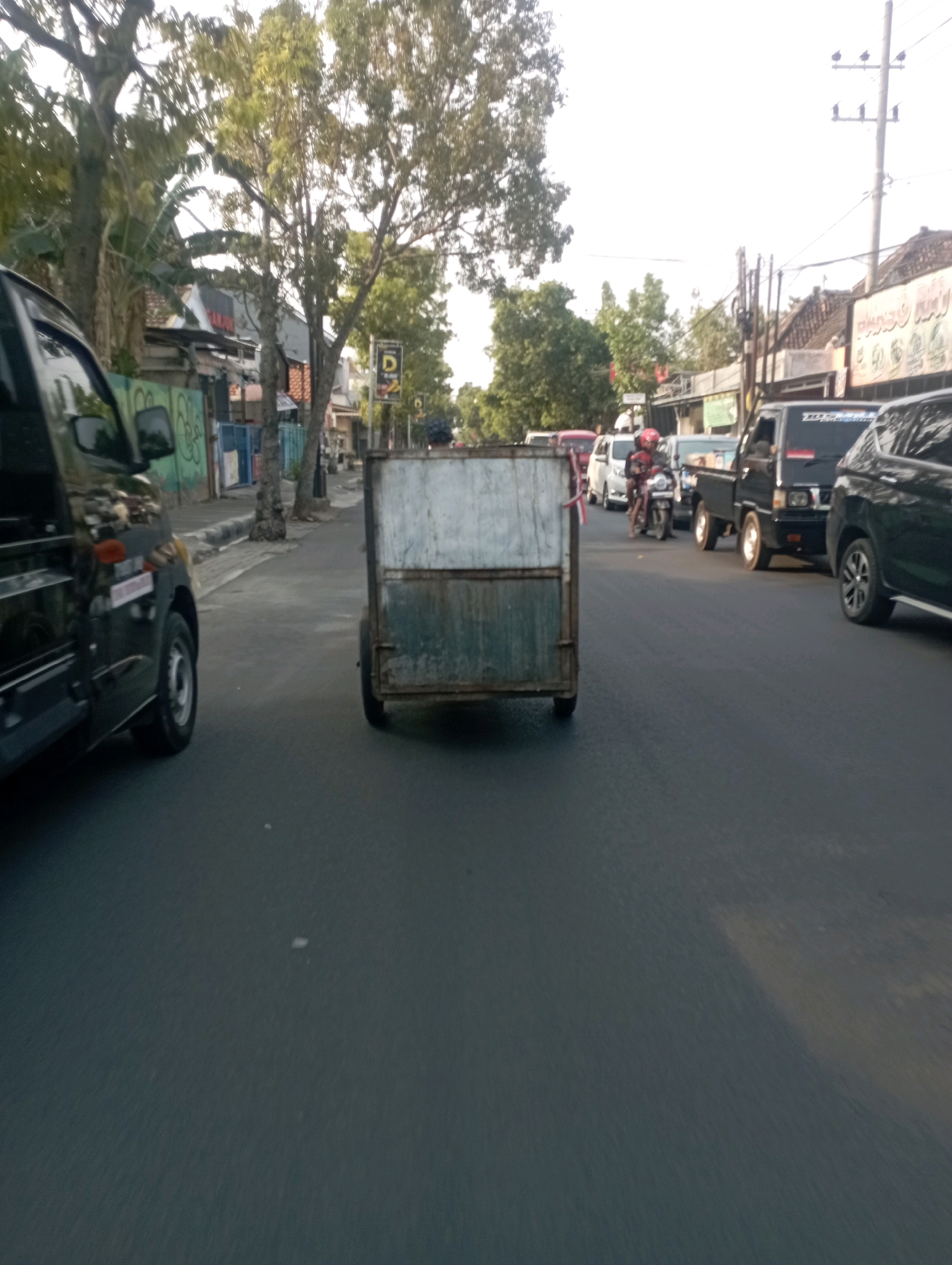 A wide-angle, low-angle shot captures a street scene in Ganung Kidul, Indonesia, under a bright, hazy sky. In the foreground, a black SUV is partially visible on the left, with its side mirror reflecting the scene. To the right, a two-wheeled cart with a weathered, possibly metal, enclosure is positioned in the center of the road. The cart has a rusty appearance, with green and white panels.
Further down the road, traffic is moderately heavy, featuring several vehicles including a black pickup truck with yellow accents on its wheels and a white minivan. A person wearing a red helmet is riding a motorcycle in the lane ahead of the cart. The street is lined with trees and buildings, suggesting a commercial or residential area. A utility pole with multiple crossbars and insulators stands on the right side of the road, further down. Signs are visible on some of the buildings, though they are not clearly legible. The overall impression is of a busy street during daytime.