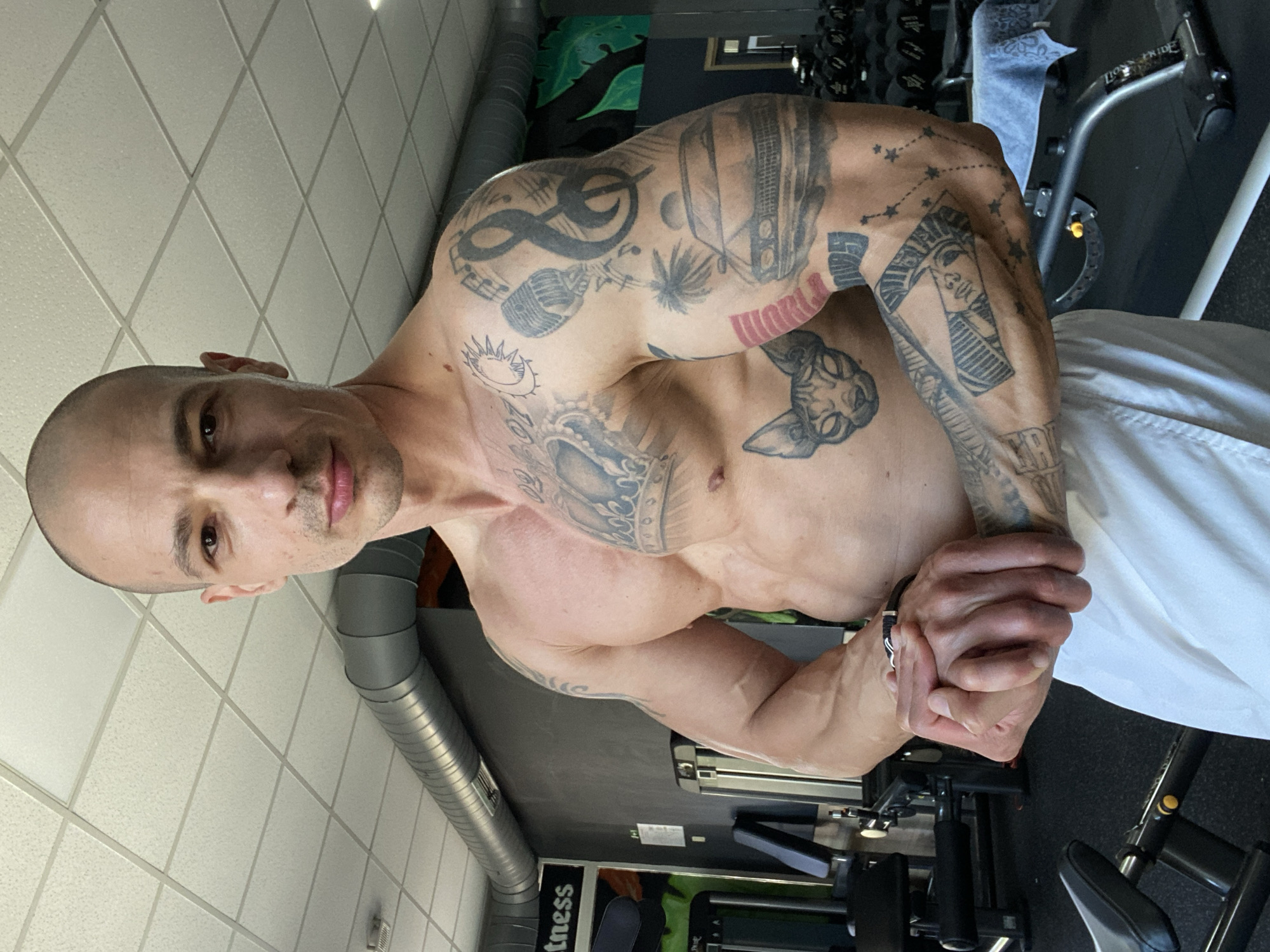 A bald, light-skinned man with tattoos is posing in what appears to be a gym setting. The man is shirtless, flexing his arms and upper body muscles. His arms and shoulders are extensively tattooed with a variety of designs. He is wearing white pants or shorts. The background includes gym equipment, such as dumbbells and a weight machine. A ventilation duct runs along the ceiling of the room. "fitness" is visible on the gym equipment. The location of the image cannot be confirmed from the image provided.