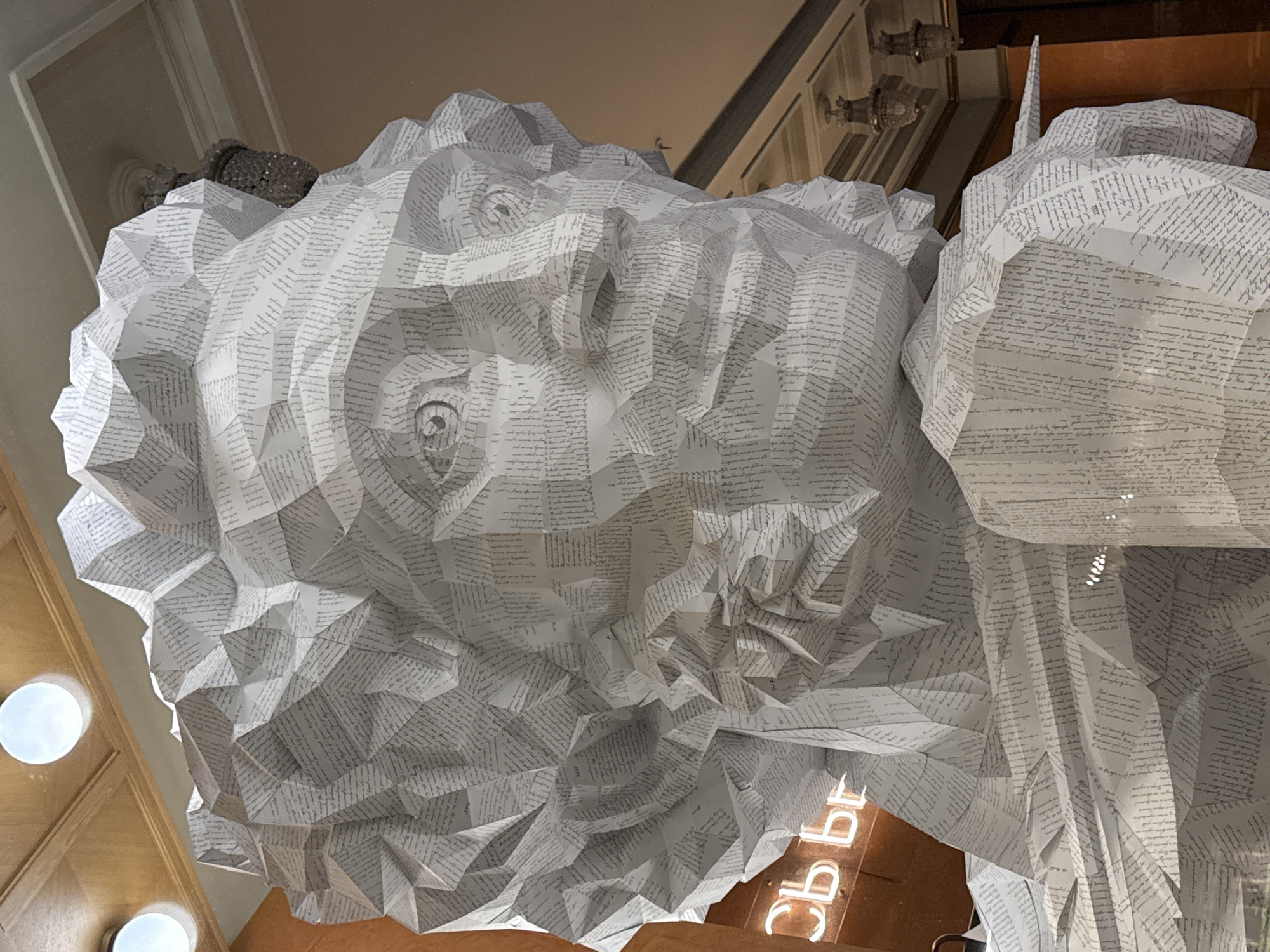 A large, intricate polygonal sculpture of a male bust dominates the foreground, crafted entirely from white paper sheets printed with dense gray Cyrillic text. The sculpture depicts a man with curly hair, distinct facial features including eyes, nose, and mouth, all rendered with sharp, angular facets. The bust extends to the upper chest, with a pointed element protruding from the right shoulder, resembling a quill or pen.
The scene is set indoors, within an ornate building in Saint Petersburg, Russia. Overhead, a light-brown wooden ceiling with decorative trim features several circular, recessed lights emitting a soft white glow. The background wall is a muted beige or cream color. To the left, a luminous orange-red neon sign displays the Cyrillic characters "Сь вг", partially obscured, suggesting a longer word or abbreviation. To the right, two elaborate chandeliers with crystal elements hang from the high ceiling, contributing to the classical interior aesthetic. The lighting is generally even, highlighting the texture and folds of the paper sculpture. The sculpture's material and style, combined with the context of Saint Petersburg and the ornate interior, strongly suggest it is the "Paper Pushkin" installation located within the Dom Knigi (House of Books) on Nevsky Prospekt.