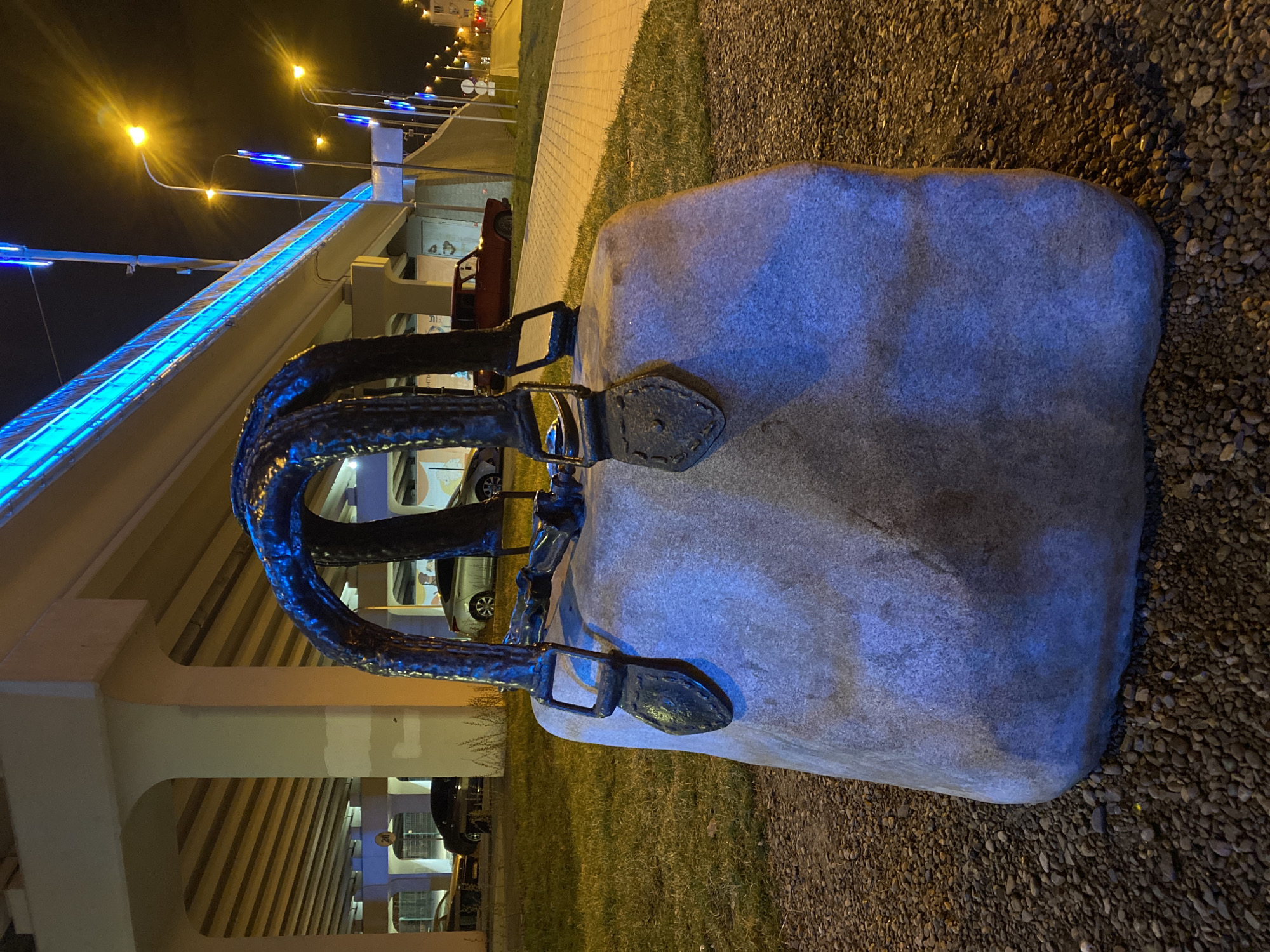 The image captures a suitcase monument at night in Yekaterinburg, Russia. The monument is composed of a large, roughly rectangular stone with an attached metal handle and buckles, shaped to resemble a vintage suitcase. The monument sits on an area covered with small, dark gravel.
In the background, a brightly lit overpass structure is visible, illuminated with blue and yellow lights. The overpass columns are white. Underneath the overpass, cars are parked. Streetlights are visible in the distance. Some green grass is seen between the suitcase monument and the overpass. The sky is dark.