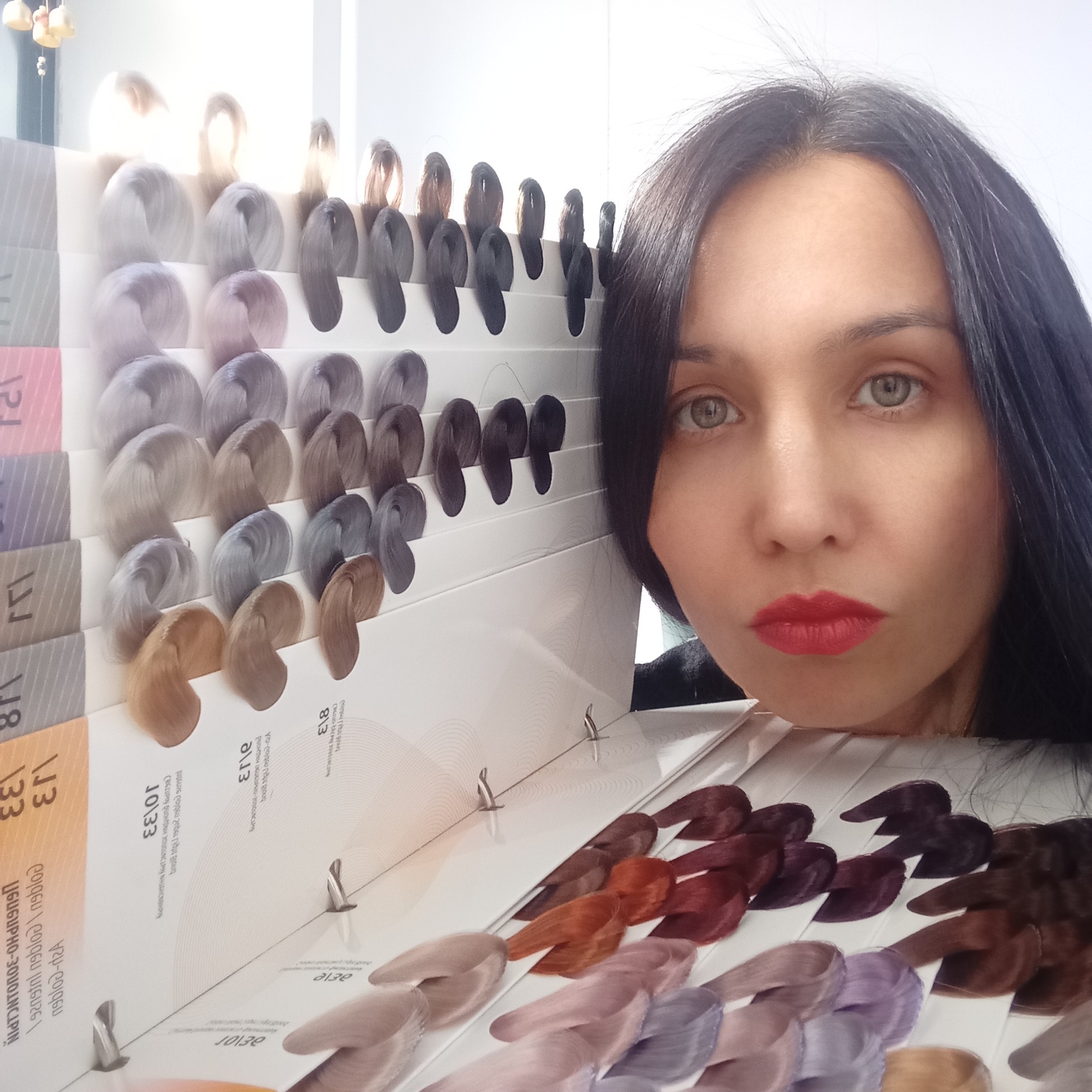The image captures a close-up of a woman next to an open hair color swatch book in what appears to be an indoor setting, likely a hair salon, in Izhevsk, Russia. The woman, with dark hair, fair skin, light eyes (green/hazel), and vibrant red lipstick, gazes directly at the camera. Her head and shoulders are visible, partially obscured by the large hair color chart she is holding or leaning against.
The main object is a comprehensive hair color chart, featuring numerous rows of synthetic hair swatches arranged by shade. A wide spectrum of colors is displayed, including various shades of blonde, ash, grey, brown, red, and purple/lavender tones. Many swatches are attached to white cards that bear printed numbers and text, some of which appear mirrored or upside down due to the angle of the photograph. The overall lighting is bright, possibly from an overhead source or a window, causing some glare on the upper portion of the chart.
The activity depicted is likely the process of selecting a hair color, either by a client or being presented by a professional.
Visible text on the cards includes numbers such as "118 33", "10130", "10\33", "813", and "8/1". Some words like "Ash" and "Golden" can be discerned, along with potentially garbled or stylized brand names like "GUGUPHO-3000INCIPIN", "PROHIINH UGUGUPNG-ONCU", and "nablo-".