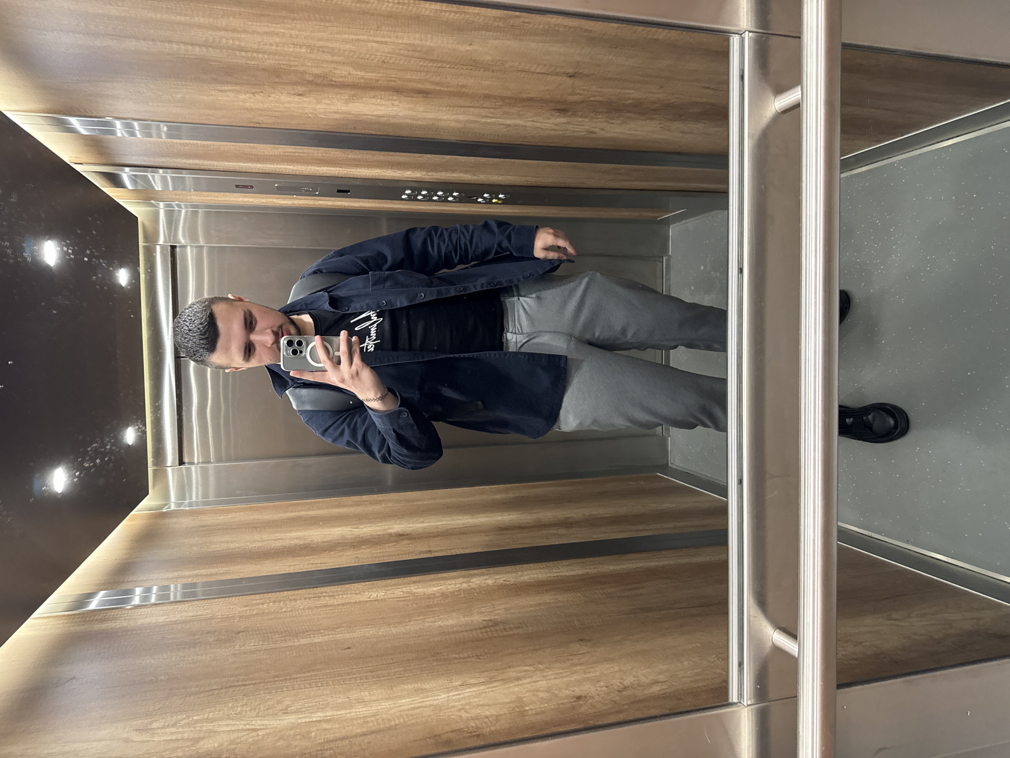 A man is taking a selfie in an elevator with wood paneled walls on three sides and a grey floor. He is wearing grey pants, a black T-shirt, and a blue jacket. The elevator has metal trim and control panels. The specific location in Sofia, Bulgaria, is not discernible from the image.