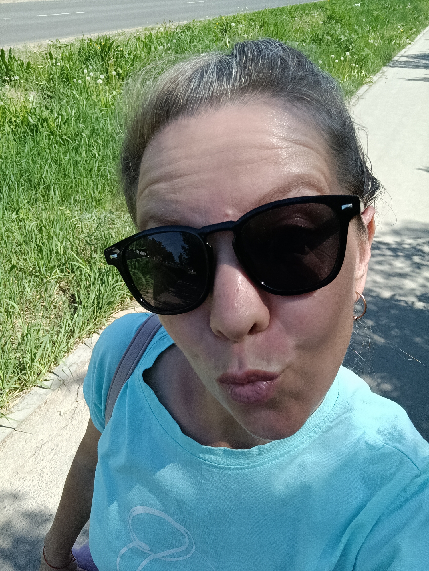 A medium-close-up, high-angle, full-daylight photograph depicts a woman in an outdoor setting. She is fair-skinned, wearing black sunglasses, a light blue t-shirt, and hoop earrings. The woman's hair is pulled back. She is looking into the camera with pursed lips. Over her left shoulder is a pale pink strap.
The background includes green grass to the left and a light-colored concrete walkway to the right. Beyond the grass is a paved road with sparse vegetation along the side. Shade falls on the right side of the walkway. The reflection in the sunglasses suggests a street with trees and possibly buildings. The location is possibly in the city of Chaikovsky, Russia.