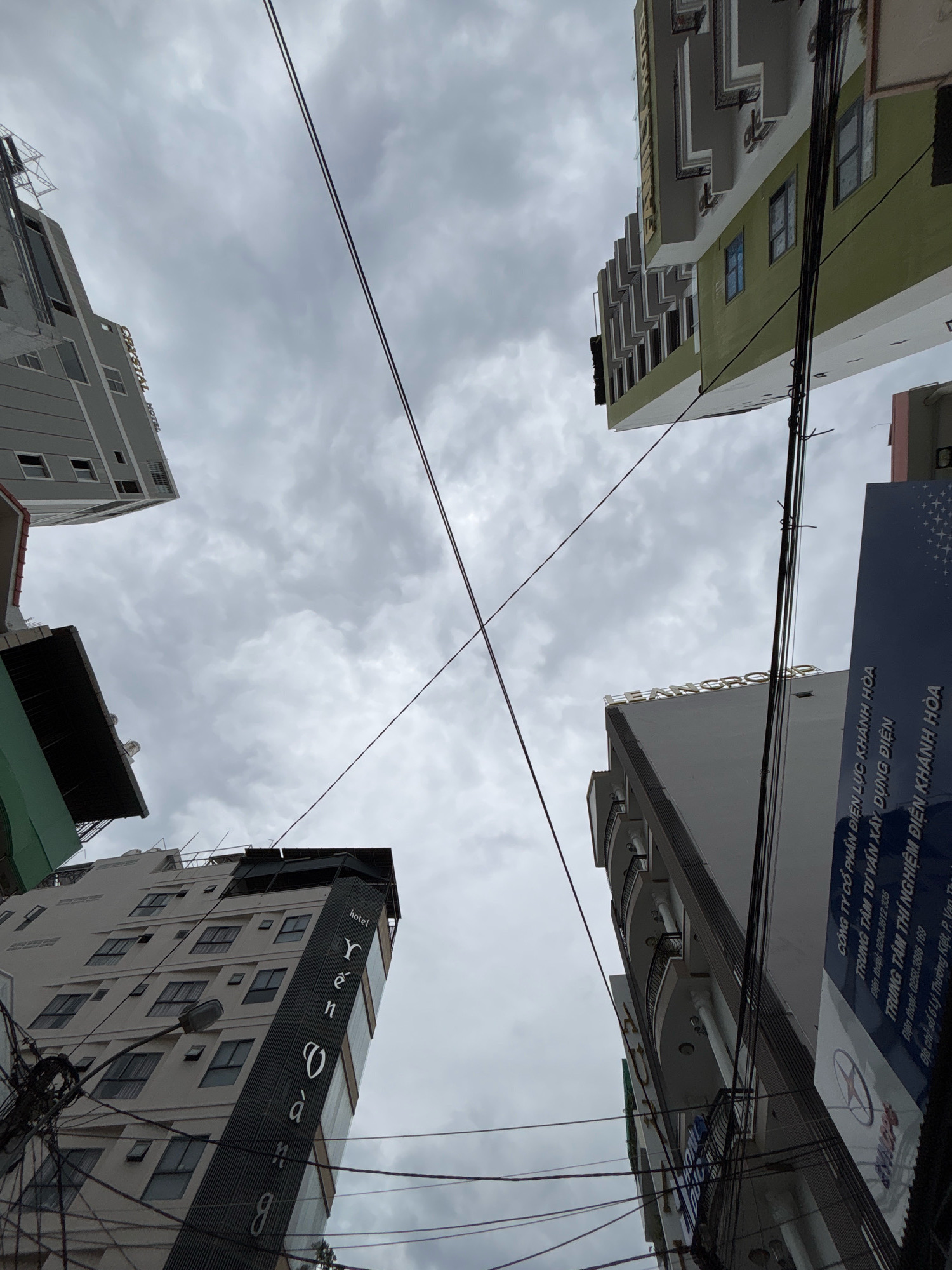 The image is a low-angle, wide shot of several buildings in Nha Trang, Vietnam, against a cloudy sky. The buildings are oriented at various angles, creating a sense of depth and urban density. In the foreground, a tall, light-colored building with dark accents features the vertical text "Yến Sào" and the word "hotel" in smaller font. This building appears to be a hotel or a business related to bird's nest products, a common commodity in Vietnam. To the left, another building, partially obscured, has the word "CRYSTAL HOTEL" visible vertically. On the right, a dark blue sign with white text is attached to a building, displaying company information and contact details, including "TRUNG TÂM TƯ VẤN XÂY DỰNG ĐIỆN" and a phone number. Overhead, a tangle of electrical wires crisscrosses the frame, a common sight in Vietnamese urban landscapes. The architecture of the buildings varies, with some featuring balconies and modern facades, while others appear older or under construction. The overall atmosphere is overcast and somewhat dim due to the cloudy weather.
