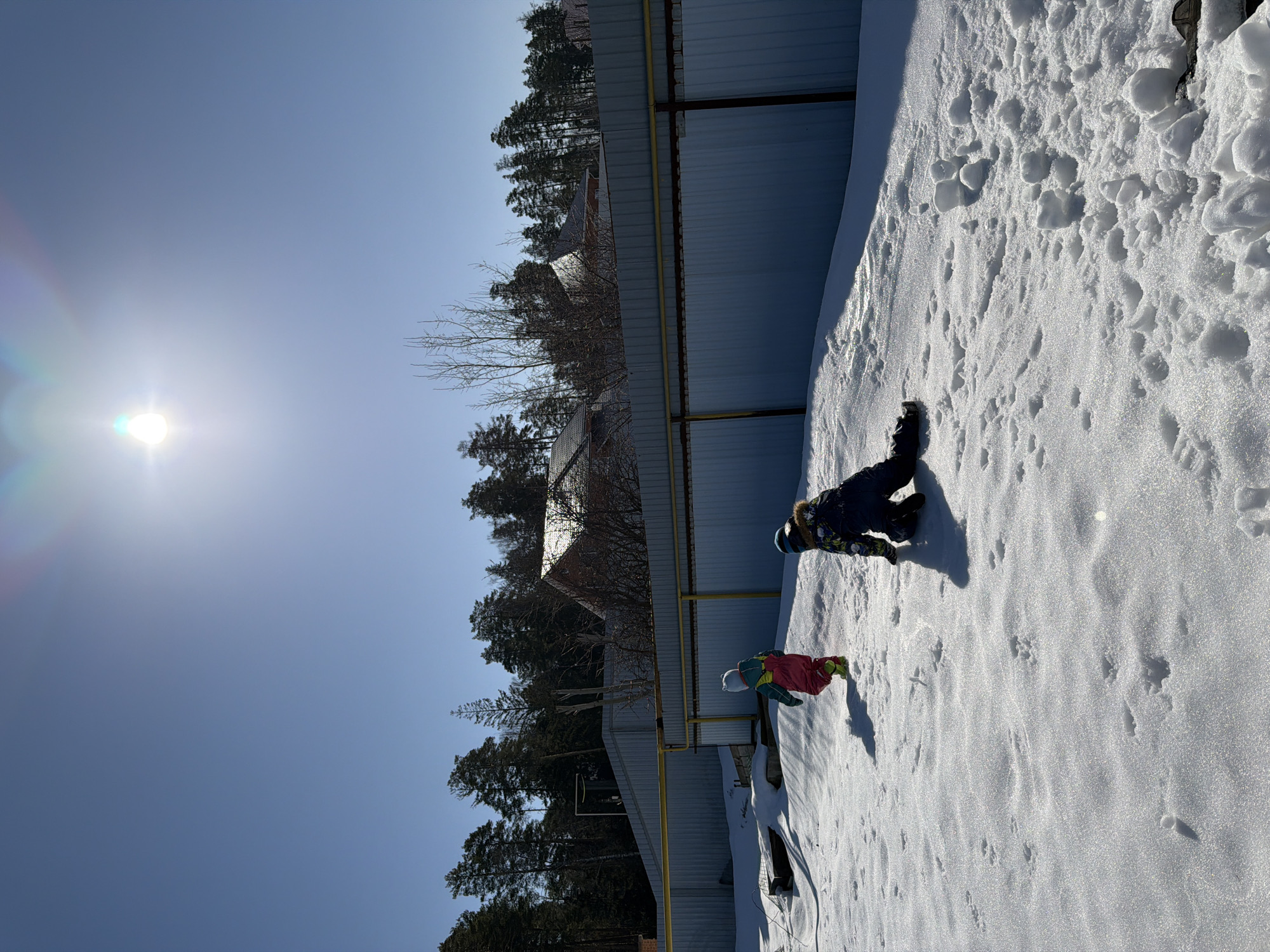 The image depicts a snowy landscape in what appears to be a backyard or open area near a residential zone in Chaikovsky, Russia. A metal fence runs along the scene, with trees visible in the background. A child wearing winter clothing is lying in the snow. A sled or another child is next to them. The scene suggests winter activities in a residential area with snow cover. The bright sunlight indicates a clear day.
