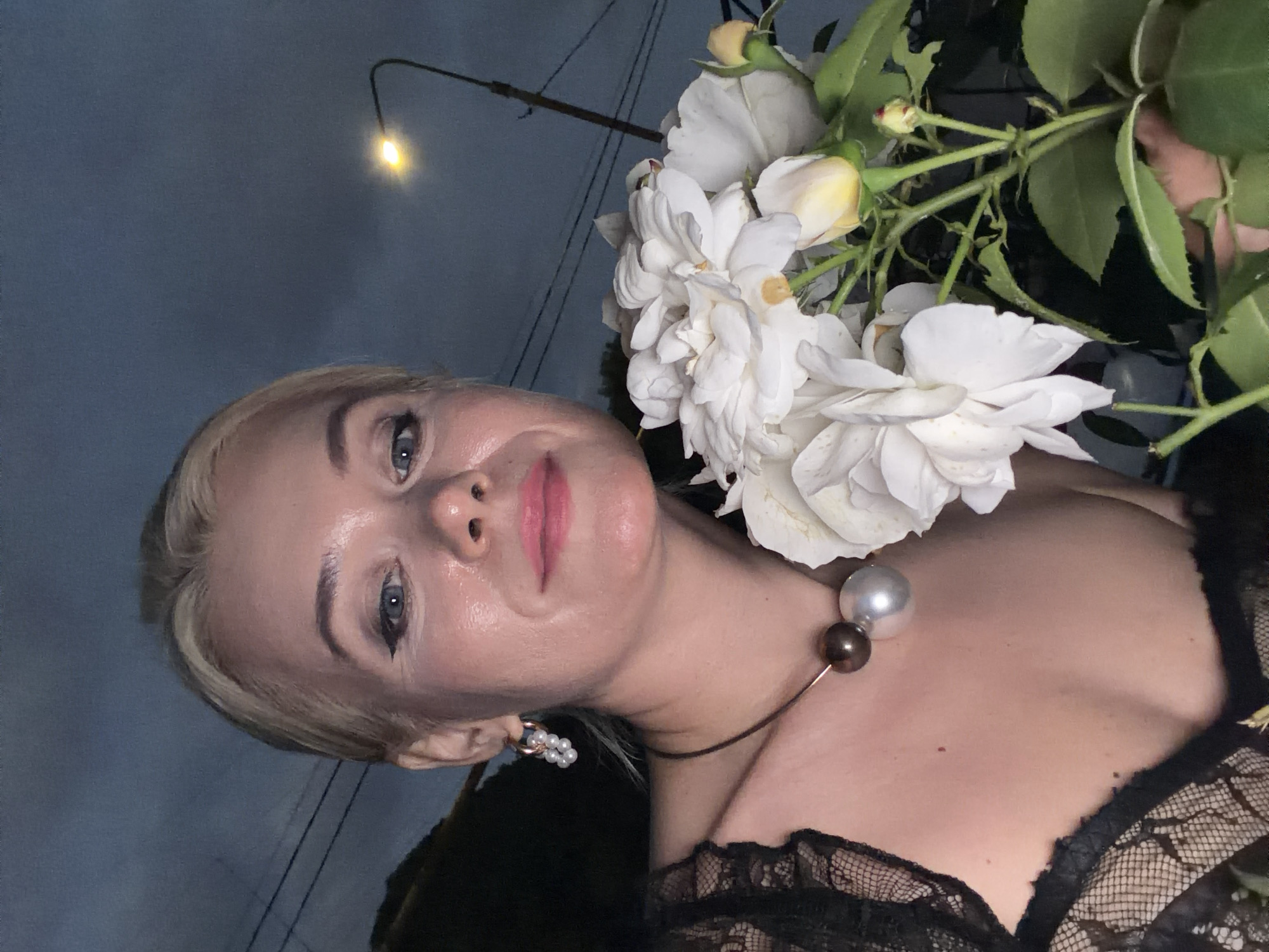 A woman with blonde hair and blue eyes is shown from the chest up, with her head tilted. She is wearing a black lace top and a necklace with two large beads, one silver and one brown. She also has pearl earrings. She is holding a bouquet of white roses. The background is a dark, blurry sky with a single street lamp visible. The lighting suggests it is nighttime. The overall mood is elegant and sophisticated. The location context is Sochi, Russia. There is no visible text in the image.