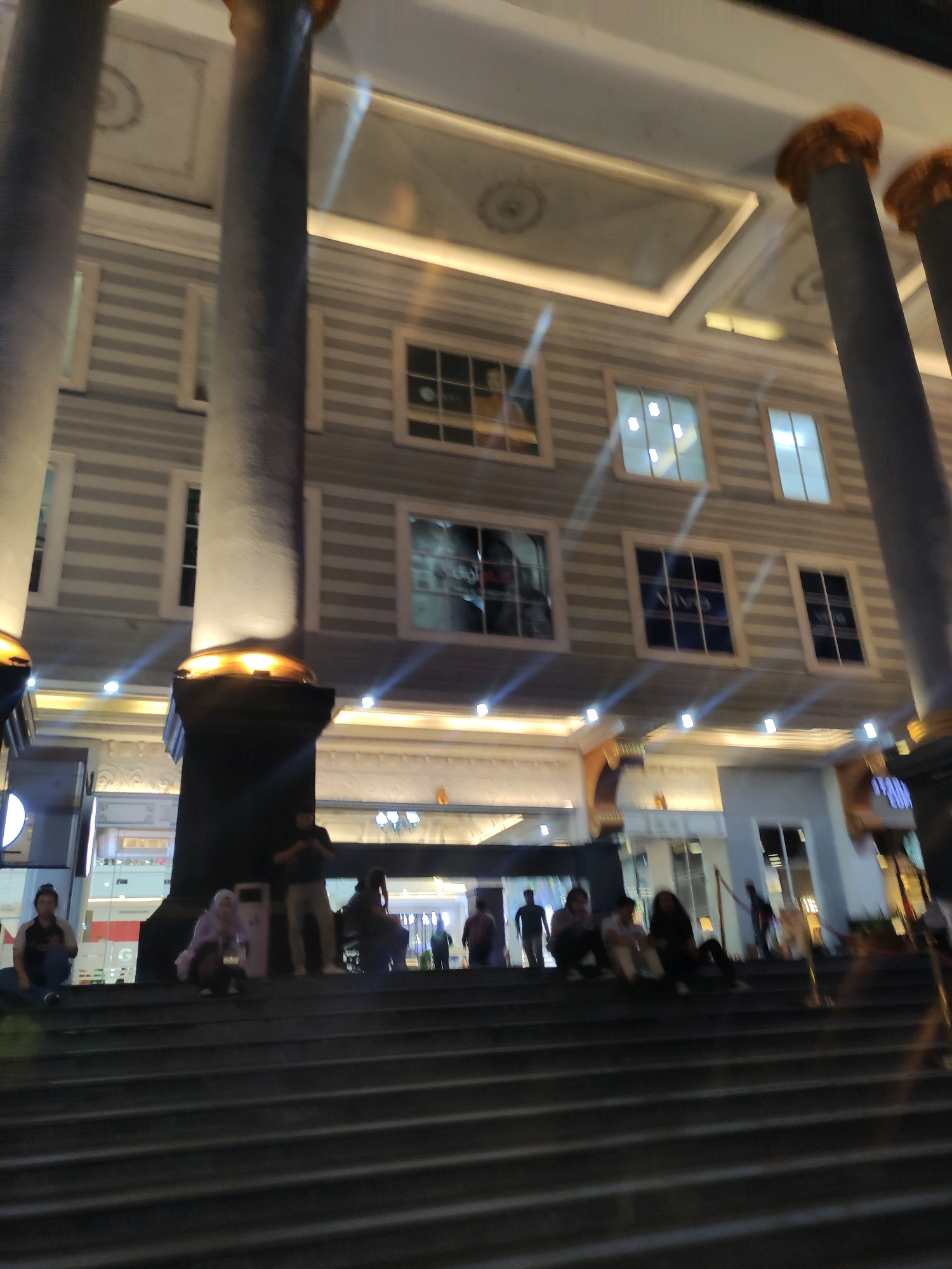 This is a nighttime, eye-level, slightly tilted full shot of the exterior of a building with a grand architectural style. The building features large, dark columns with golden tops, a striped facade, and multiple rows of illuminated windows. Below the building, a wide set of dark stairs descends into the foreground, occupied by several individuals sitting or standing. Some people are facing the camera, while others are engaged in conversation or looking away. The lighting on the building is a mix of warm yellow from decorative lights at the base of the columns and brighter white lights from the windows, creating a vibrant and somewhat ethereal atmosphere. In the background, more of the building's facade and illuminated entrances are visible, suggesting it is a commercial or entertainment venue. The overall scene depicts a lively public space at night. The location is Sinduadi, Indonesia.