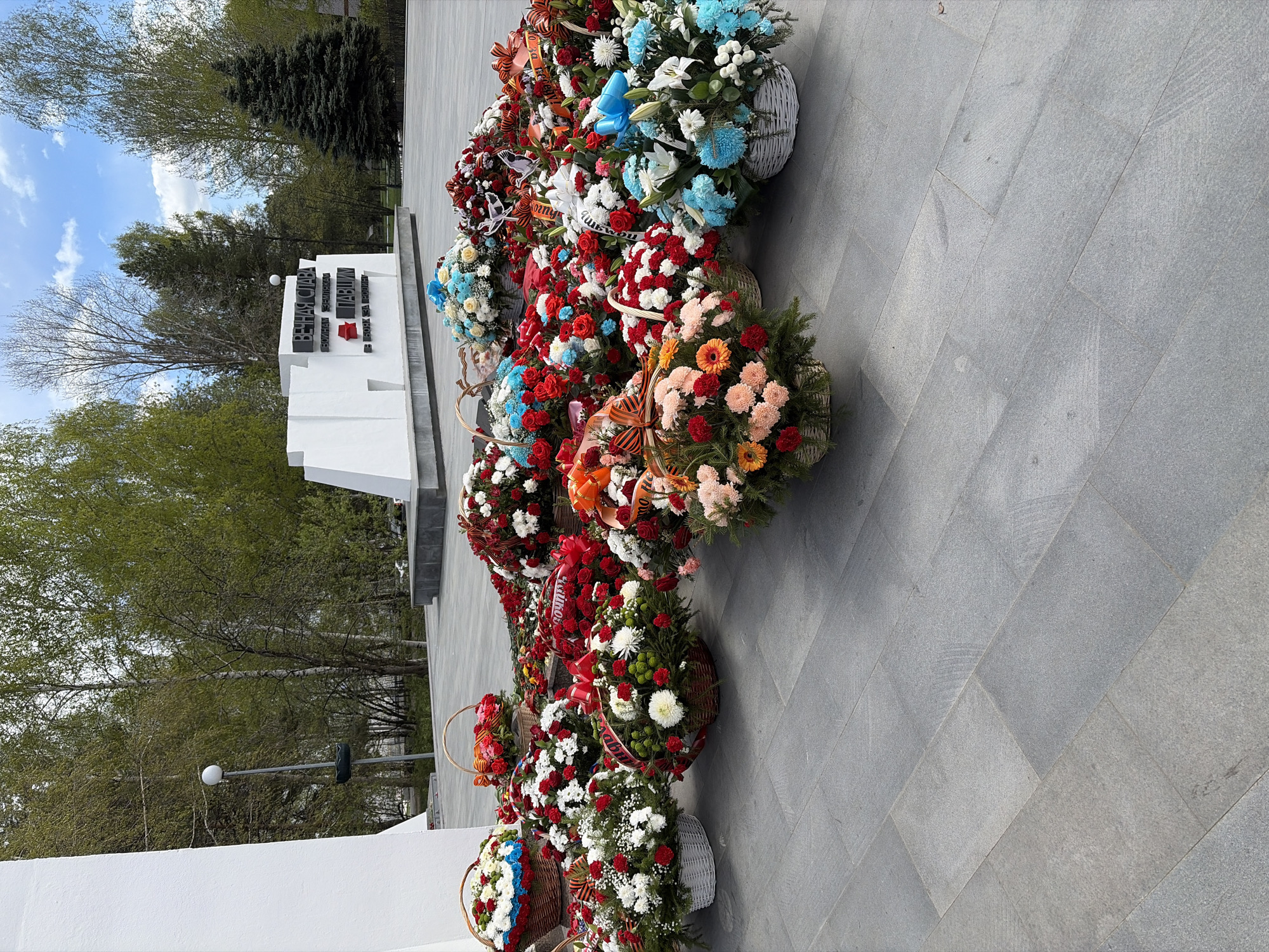 The image shows a memorial with a white structure and dark lettering, possibly a war memorial. In front of the memorial, there are multiple flower baskets in a row. The baskets contain a mix of flowers in red, white, blue, and orange colors. The memorial is located in an outdoor setting. Trees can be seen in the background. The ground is paved with grey tiles. The image was taken in the city of Chaikovsky, Russia.