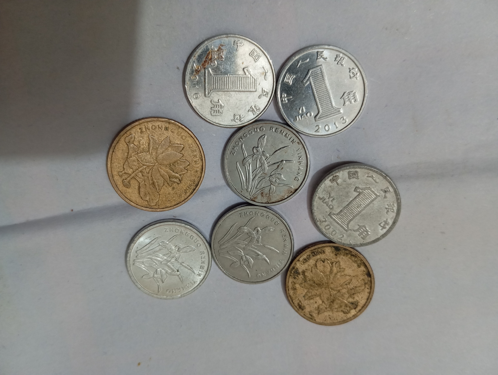 Eight coins are scattered on a neutral gray surface. The coins are a mix of silver and brass colored. The silver coins appear to be from China, with inscriptions in Chinese characters and Arabic numerals. Some of the silver coins feature the number "1" prominently, along with the text "JIAO" and years such as "2013" and "2002." Other silver coins display images of plants, specifically what appear to be orchids, and the text "ZHONGGUO RENMIN YINHANG." The brass-colored coins also feature plant imagery, with one showing a lotus flower and another coin, partially obscured, showing a similar floral design. The coins are not arranged in any discernible pattern and are spread across the frame, with some overlapping slightly. The lighting suggests an overhead source, casting faint shadows. The scene is devoid of any other objects or subjects.