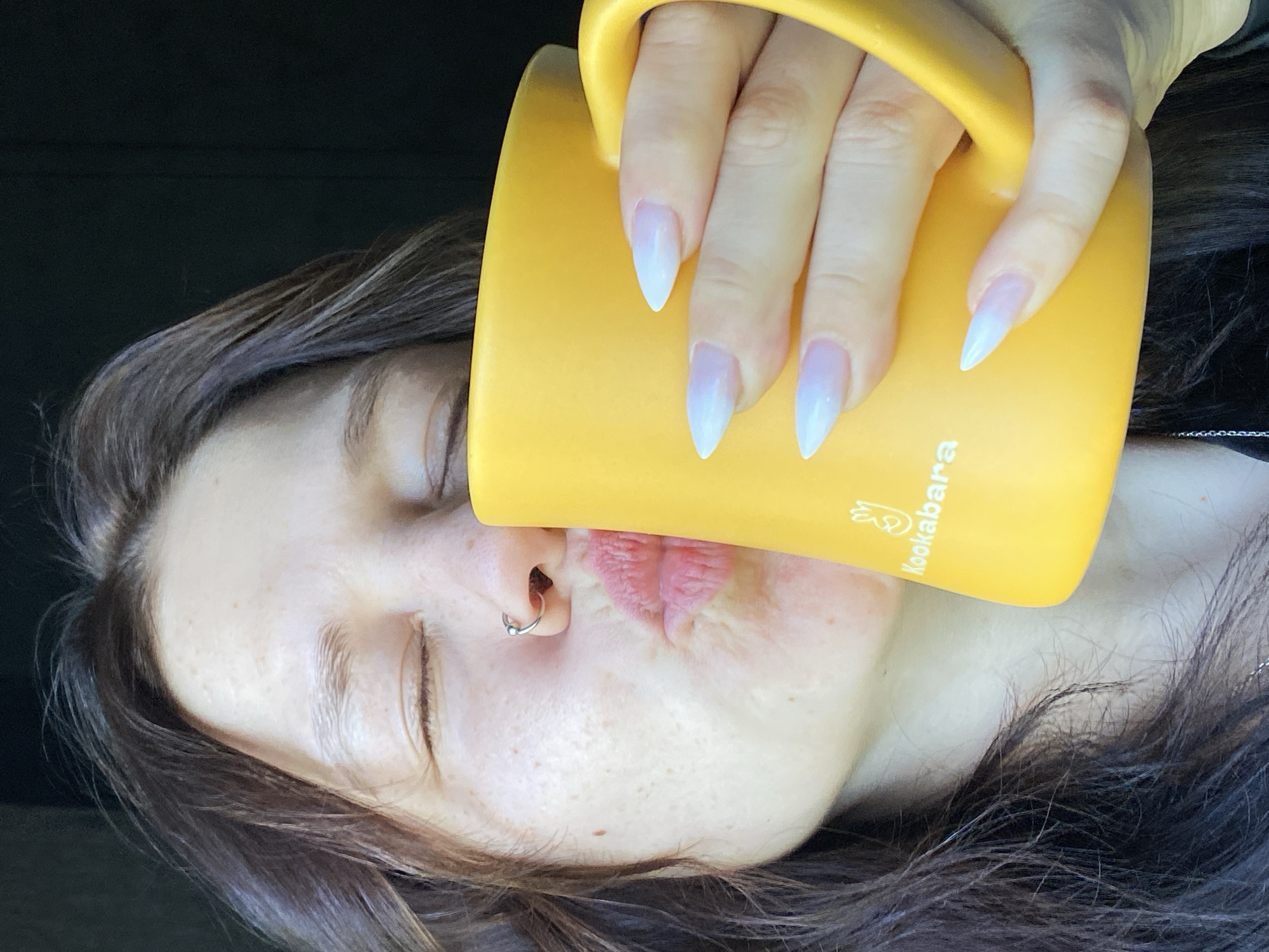 A close-up, horizontally oriented image captures a person taking a drink from a vibrant yellow mug. The person's face is visible, with closed eyes and lips puckered around the mug's rim, indicating they are actively drinking. A small, silver hoop nose ring is worn in their left nostril. Their skin appears fair with some scattered freckles.
The person's right hand, adorned with long, pointed nails featuring a white or pale pink ombre design, holds the bright yellow mug. The word "Kookabara" is clearly visible on the side of the mug, alongside a small, stylized graphic that resembles a bird. Dark brown hair frames the person's face. The background is indistinct and dark, suggesting an indoor setting or a focused shot within a vehicle. The overall impression is one of a private moment, possibly of quiet enjoyment or refreshment, taking place during the daytime in Chaikovsky, Russia.