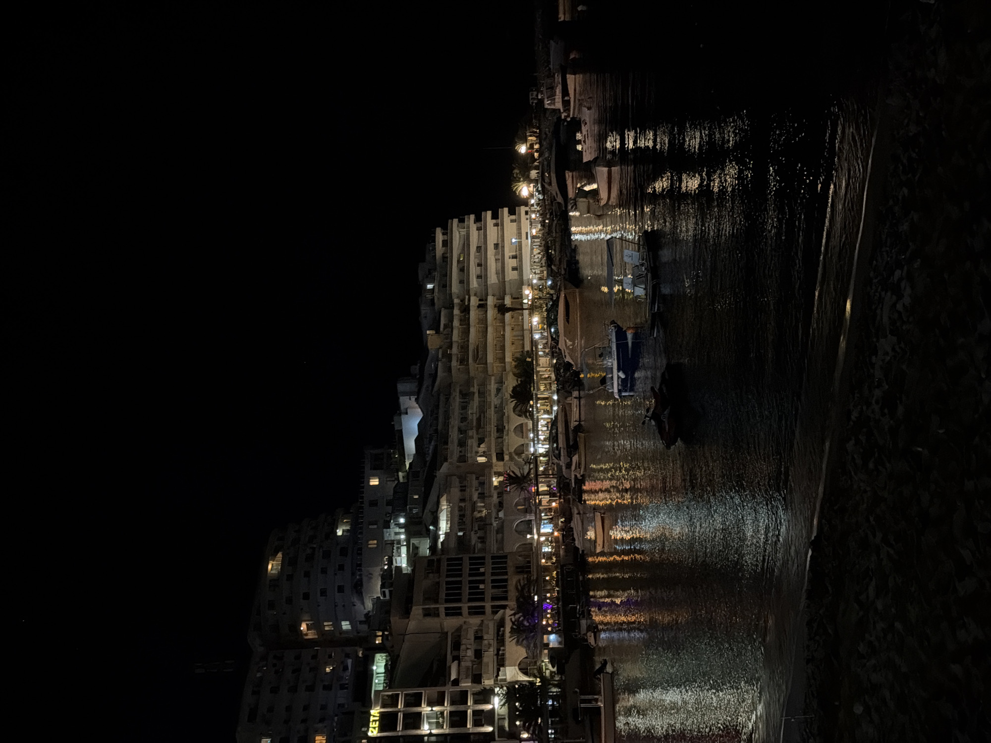 The image depicts a nighttime scene at the harbor in Boreti, Montenegro. A multi-story, curved hotel building dominates the background, illuminated by numerous lights from its windows and facade. In the foreground, the dark water of the harbor reflects the lights from the buildings and the sky, creating a shimmering effect. Several boats, including what appear to be jet skis and smaller vessels, are moored in the water. The water's edge is visible in the bottom of the frame, with dark sand forming a beach. The sky is dark, indicating nighttime, with a faint speck of light, possibly a star or distant aircraft, visible in the upper left quadrant. No people are clearly visible in the frame.