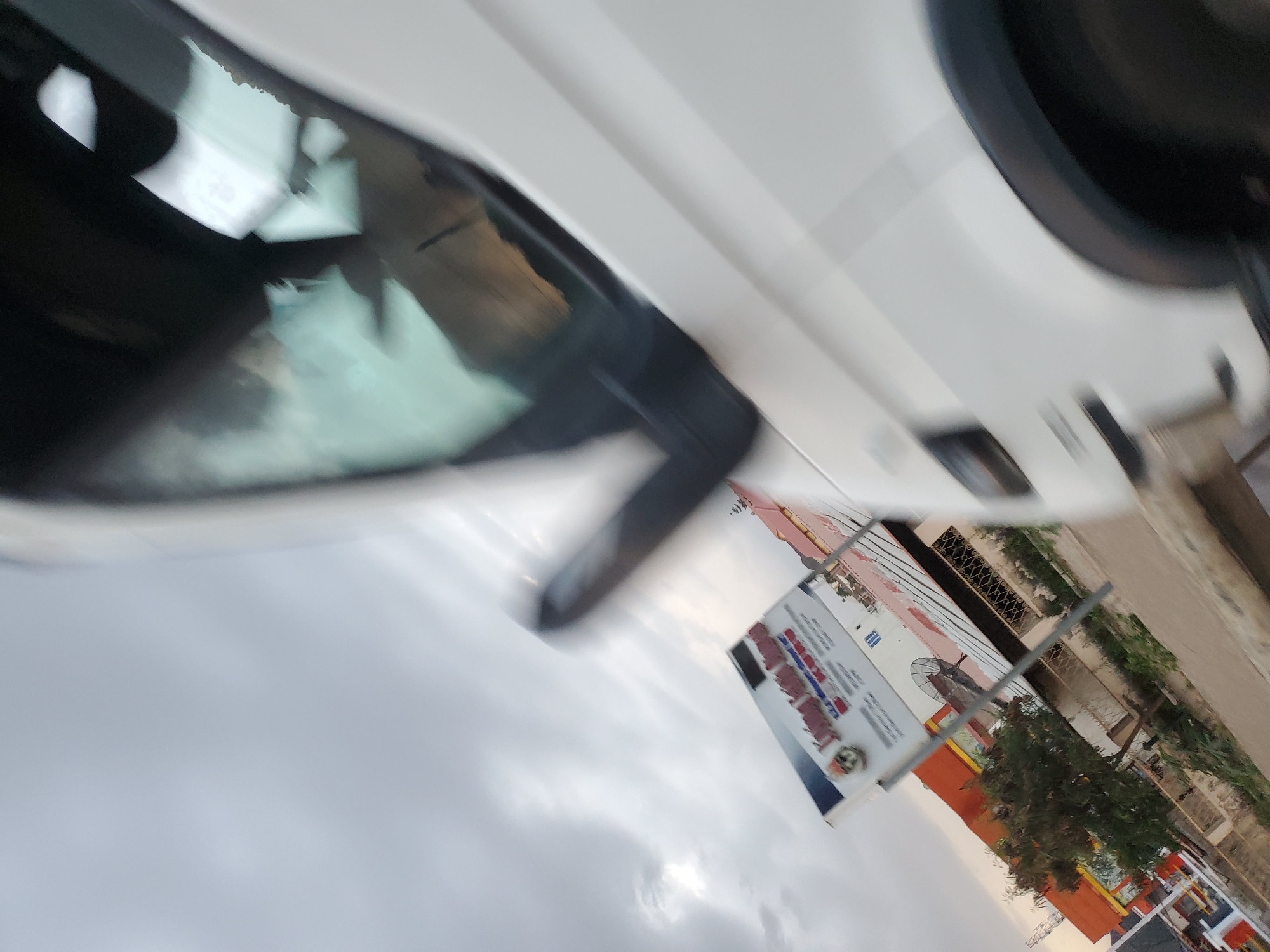 The image depicts a motion-blurred street scene in Gura Top, Nigeria. A white vehicle is prominently featured on the right side of the frame, with its side and wheel visible. In the background, a billboard with text in what appears to be a Latin script, possibly English, is mounted on poles. Below the billboard, a building with orange and white paneling is visible, along with a satellite dish and some trees. The sky is overcast and gray. The overall impression is of a roadside view captured while in motion.