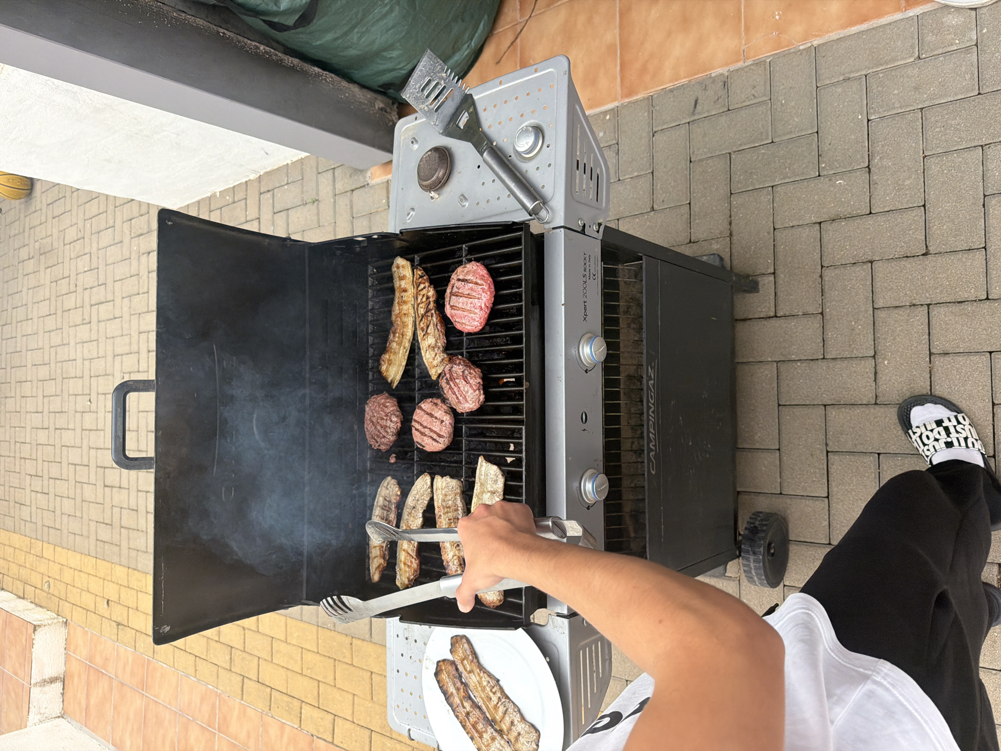 A person is grilling meat on a portable gas grill, outdoors on a brick patio, possibly in Montgó, Spain. The grill has a black lid and a silver top with a grill brush resting on it. Burgers and grilled vegetables are visible on the grill, with some already cooked on a white plate. The person is using tongs to move the food. The person is wearing black pants and a white t-shirt, with "JUST DO IT" sliders on their feet.