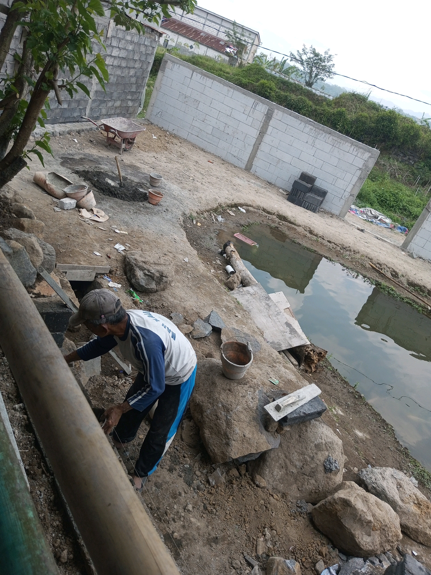 The image depicts an outdoor construction site in Kalibeber, Indonesia. A man wearing a baseball cap, a long-sleeved shirt with blue and white stripes, and dark pants is working on the ground. He appears to be using a string line and is focused on his task, with his hands near the ground. The environment is characterized by a muddy, dirt-filled area with various construction materials scattered around. Visible objects include large rocks, a wheelbarrow, concrete blocks, buckets, and a trowel resting on a large rock. A body of water, possibly a pond or canal, is present to the right of the frame, reflecting the sky. In the background, partially constructed walls made of concrete blocks are visible, along with some greenery and a building with a white roof. The overall scene suggests ongoing construction work in a rural or semi-rural setting.