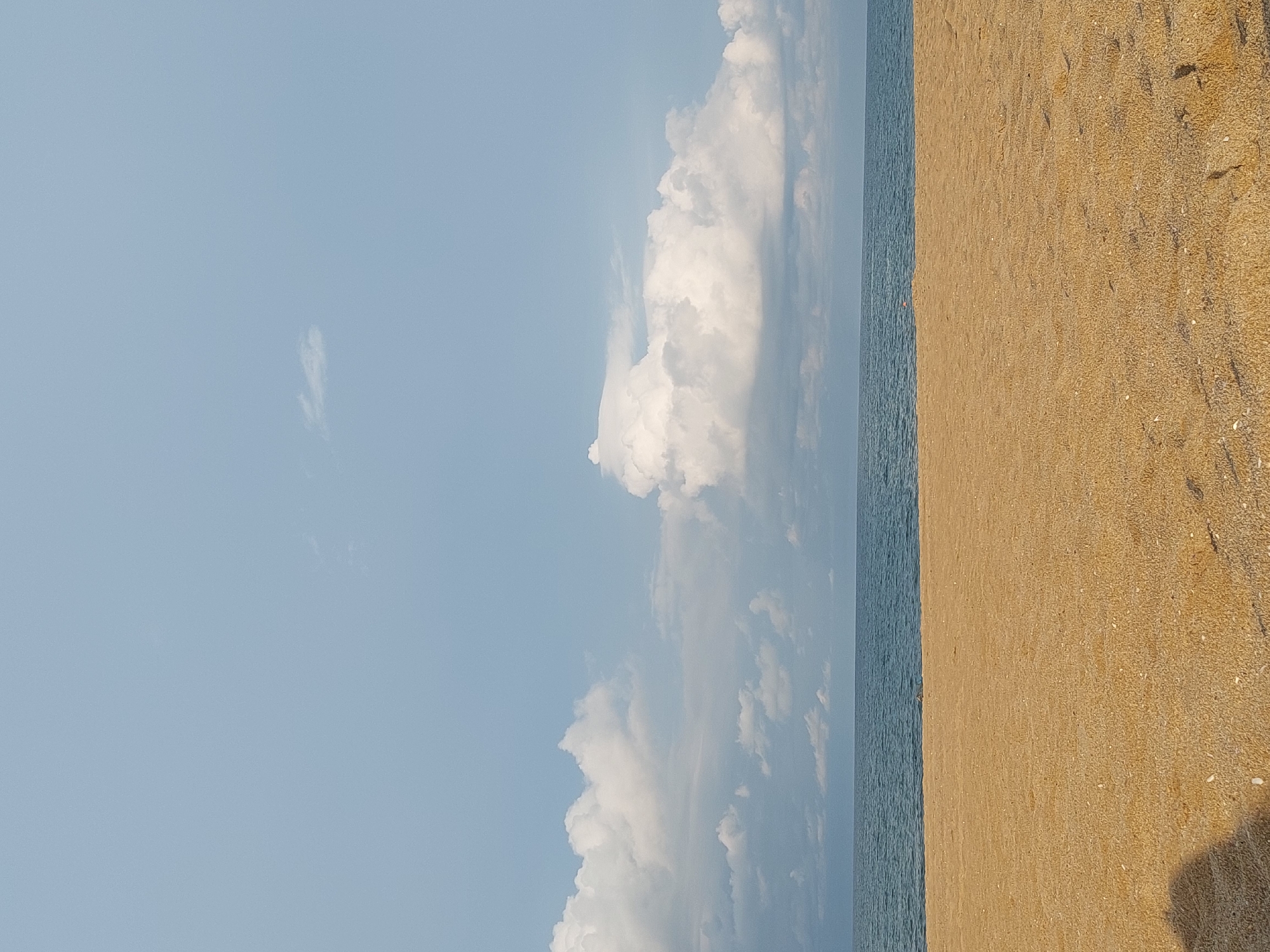 A wide-angle, eye-level shot captures a serene beach scene in Shkorpilovtsi, Bulgaria. The foreground is dominated by a vast expanse of golden sand, textured with fine grains and scattered with small shells. A dark, indistinct shadow of what appears to be a person is cast on the lower left side of the sand, suggesting the photographer's presence.
The middle ground features the calm, deep blue sea, meeting the horizon in a sharp, clear line. The water exhibits subtle ripples and is devoid of any boats or swimmers.
The upper portion of the image is filled with a pale blue sky, adorned with a variety of clouds. On the left, a line of fluffy white cumulus clouds stretches across, with some darker, more subdued clouds underneath. A prominent, large white cumulus cloud formation is centered in the frame, its edges illuminated by the sun, while others are more diffused and grey. A small, wispy cloud floats higher in the sky on the left. The overall impression is one of tranquility and natural beauty, with the focus on the expansive landscape.