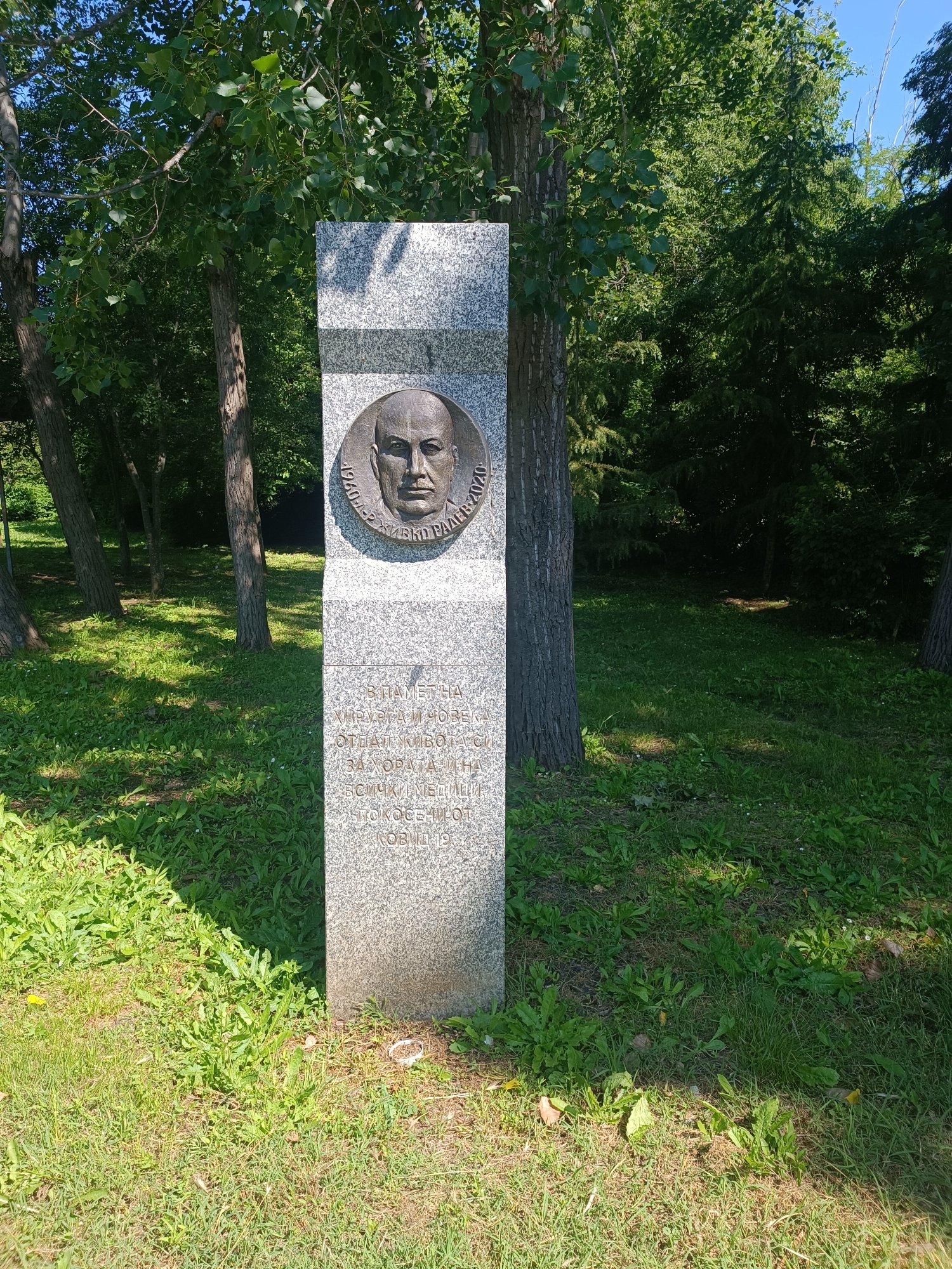 The image shows a granite memorial in a park setting. The memorial is a tall, rectangular pillar with a bronze medallion of a man's face at the top. Inscriptions are carved into the granite below the medallion. The memorial is situated on a grassy area surrounded by trees, suggesting a park or garden. The specific location in Burgas, Bulgaria, is a park dedicated to Todor Ivanchov, a Bulgarian politician and writer who served as Prime Minister. The memorial likely commemorates him or an event related to him. The date "1950-2020" is visible on the medallion, along with a name that appears to be "ТОДОР ИВАНОВ" or similar. Further inscription in Bulgarian is visible below, including the phrase "В ПАМЕТ НА ХИРУРГ И ЧОВЕК..." (In memory of a surgeon and a man...). The overall scene is one of solemn commemoration in a natural, public space.