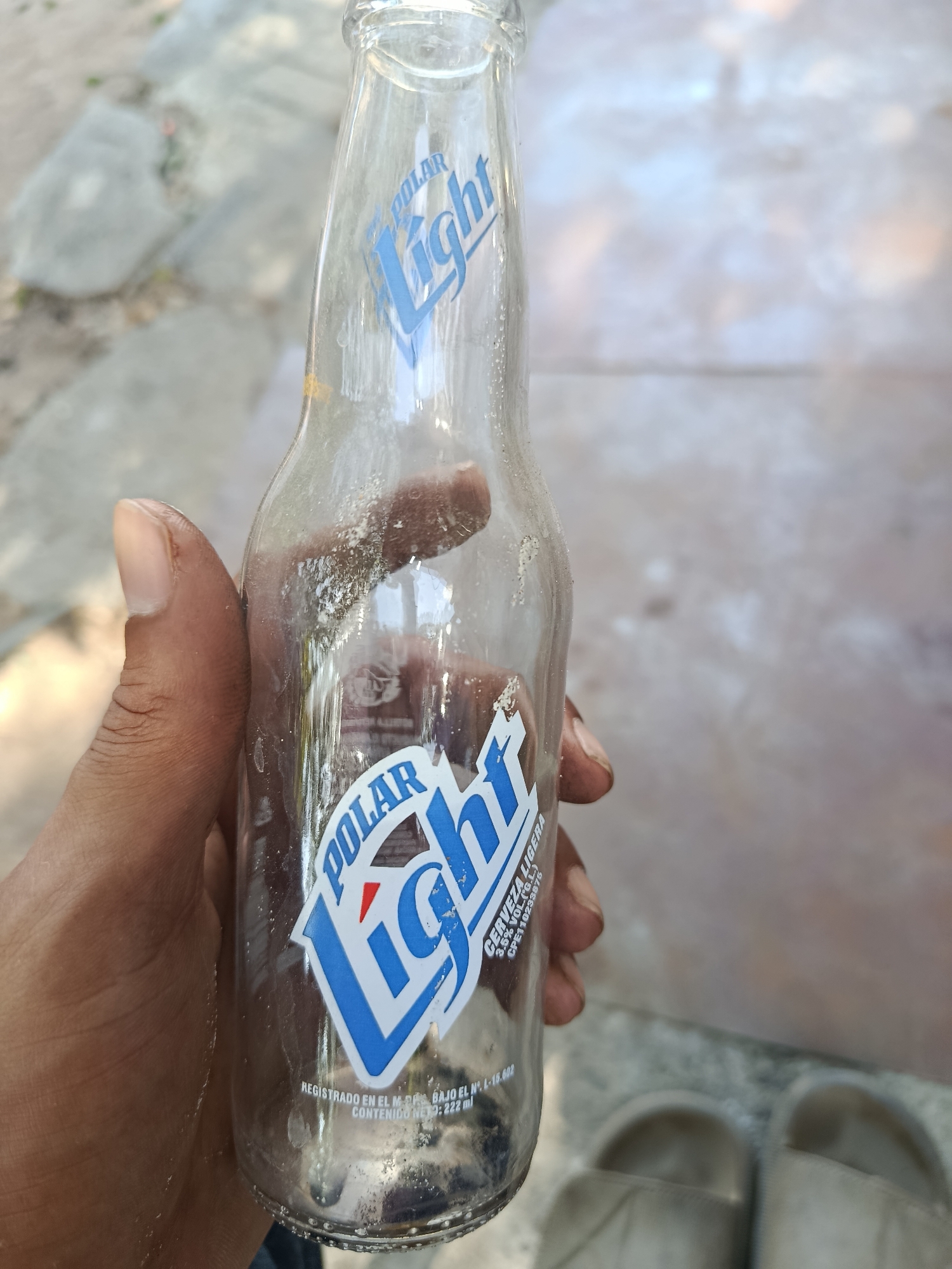 A hand holds an empty, transparent Polar Light beer bottle. The bottle shows the beer brand's logo in blue and white. The bottle is somewhat dusty and appears to have residue inside. The background shows a concrete or stone surface with some plants visible in the blurred distance. The presence of the Polar Light beer, a Venezuelan beer, implies that the photo was taken in Venezuela. Given that this beer is especially popular on Margarita Island, the photo may have been taken in Juan Griego, Venezuela.