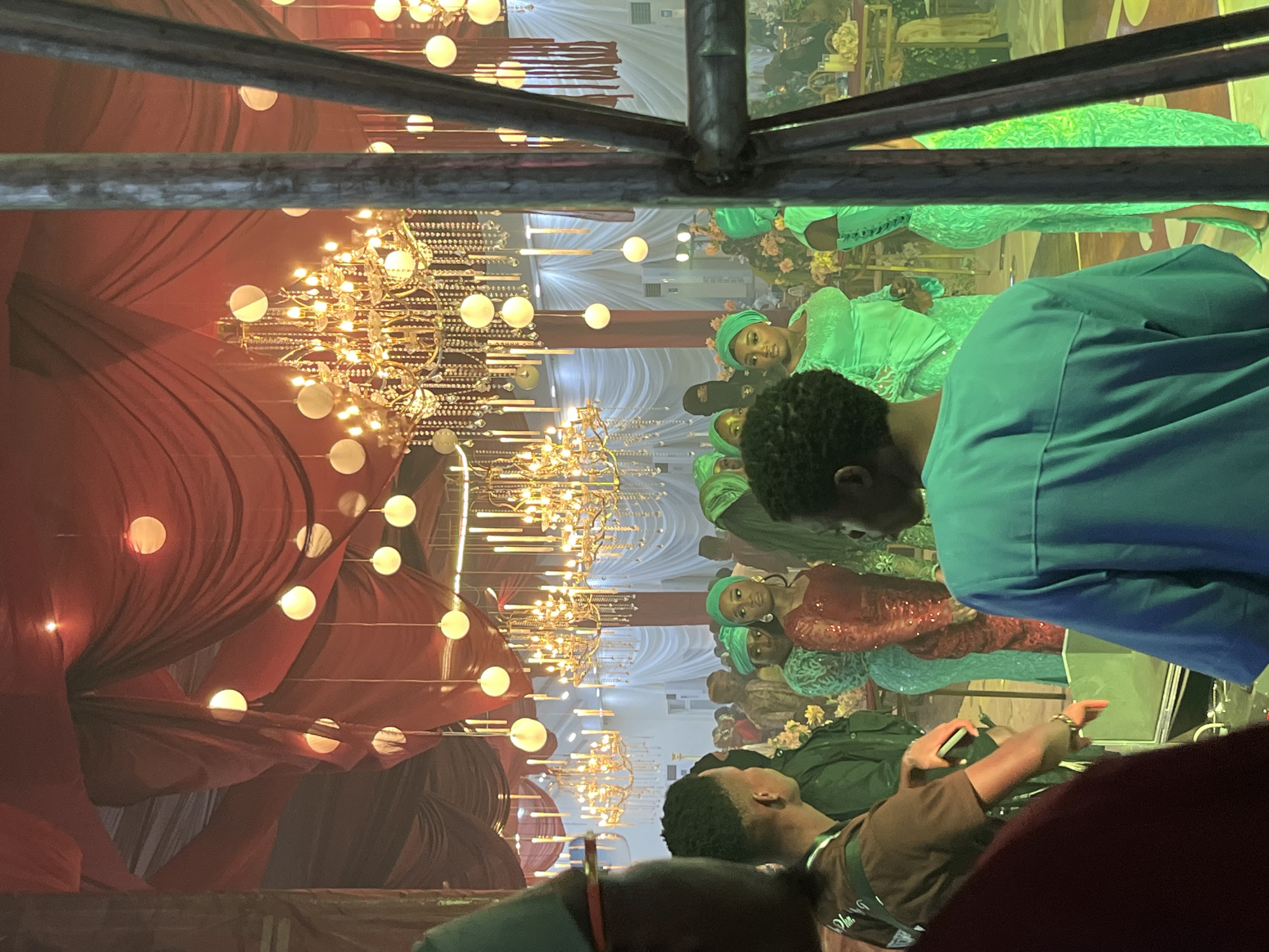 A group of people are gathered in what appears to be an indoor event space. The ceiling is draped with red fabric and adorned with several ornate chandeliers and round paper lanterns, suggesting a festive or formal occasion. In the foreground, three individuals are partially visible, with one wearing a brown shirt and another a teal blue garment. The main subjects are several women standing further back, all wearing vibrant green and red traditional attire. The women are dressed in elaborate gowns and headscarves, with some adorned in sequins. The overall ambiance is one of celebration, possibly a wedding or cultural event, with warm lighting and rich decor.
