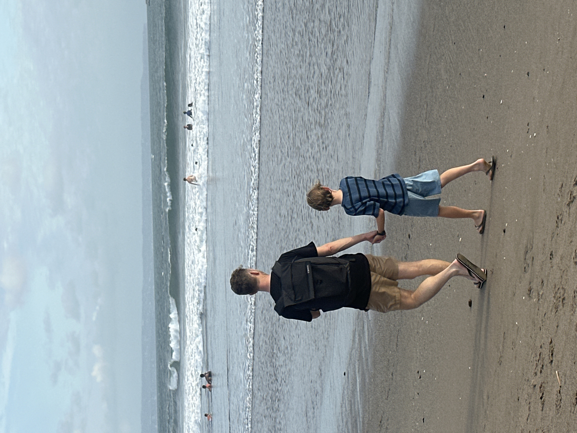 In the city of North Kuta, Indonesia, two individuals, a man and a boy, walk hand-in-hand on a sandy beach. The man, wearing a black t-shirt, khaki shorts, and flip-flops, carries a black backpack. The boy, dressed in a blue and black striped t-shirt and denim shorts, also wears flip-flops. They are walking away from the camera towards the ocean, where gentle waves are breaking. In the background, several other people are visible in the water, some appearing to be swimming and others wading. The sky is overcast with a hint of blue peeking through, and the distant horizon shows a hazy landmass. The scene captures a serene moment on a beach, likely during the late afternoon or early morning given the soft lighting.