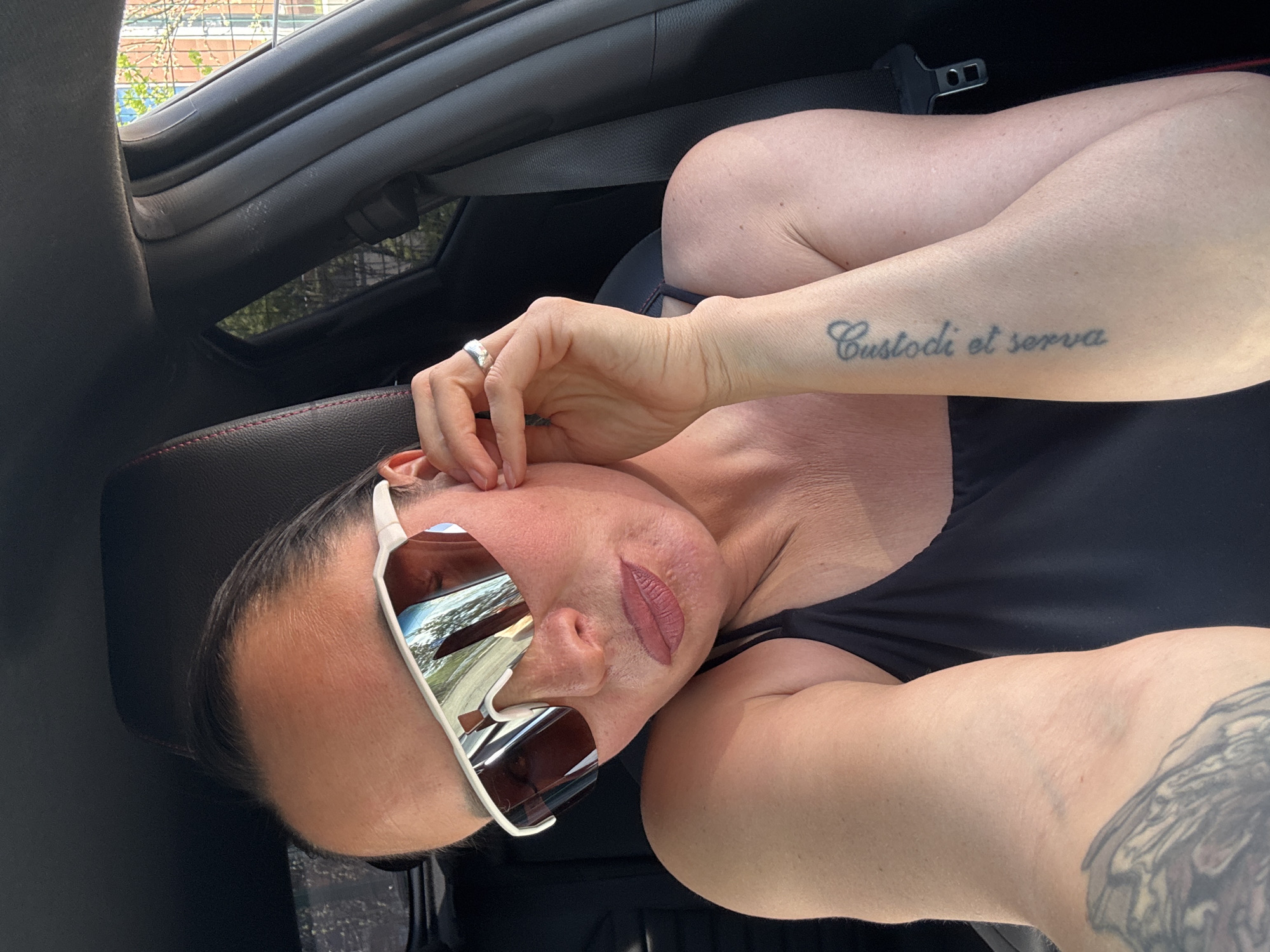 A woman is sitting in a car. She is wearing sunglasses and a black top. Her left arm is bent, and she is touching her ear with her hand. A tattoo is visible on her left forearm. There is also a tattoo on her right upper arm. A ring is on her left ring finger. The car's interior is black. The location appears to be Прикамский, Russia, based on reflections in the glasses, although this is not explicitly stated.