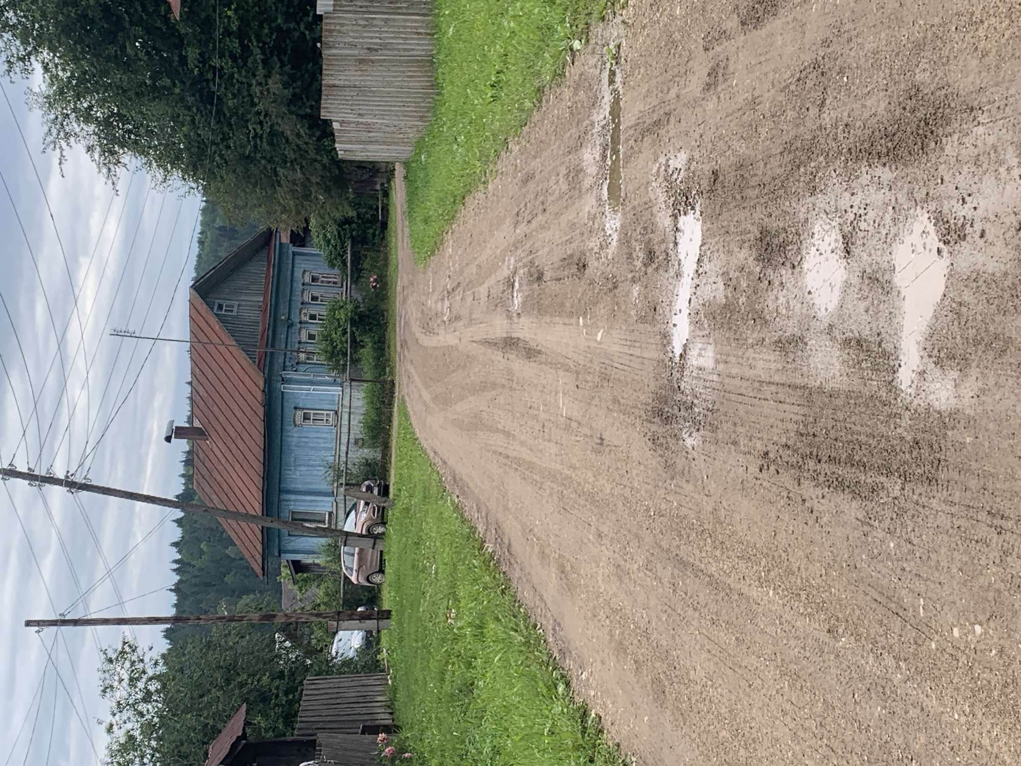 The image depicts a dirt road in a rural setting, likely in Barsai, Russia. The road is wet in places, with tire tracks visible, suggesting recent rainfall. On the left side of the road, a long, blue wooden house with a red roof stands prominently. Several windows with white trim are visible on the house. A light-colored car is parked in front of the house, and another vehicle, possibly a white van, is partially visible further down the road. Utility poles with wires stretch across the scene, indicating the presence of electricity or communication services. Dense green trees and foliage surround the house and extend up a gentle slope in the background, forming a backdrop of a wooded area. The overall impression is one of a quiet, perhaps somewhat weathered, rural landscape under an overcast sky, suggesting a daytime scene. There are no people visible in the image. The weathered appearance of the house and the presence of utility poles are consistent with a village or countryside location in Russia.