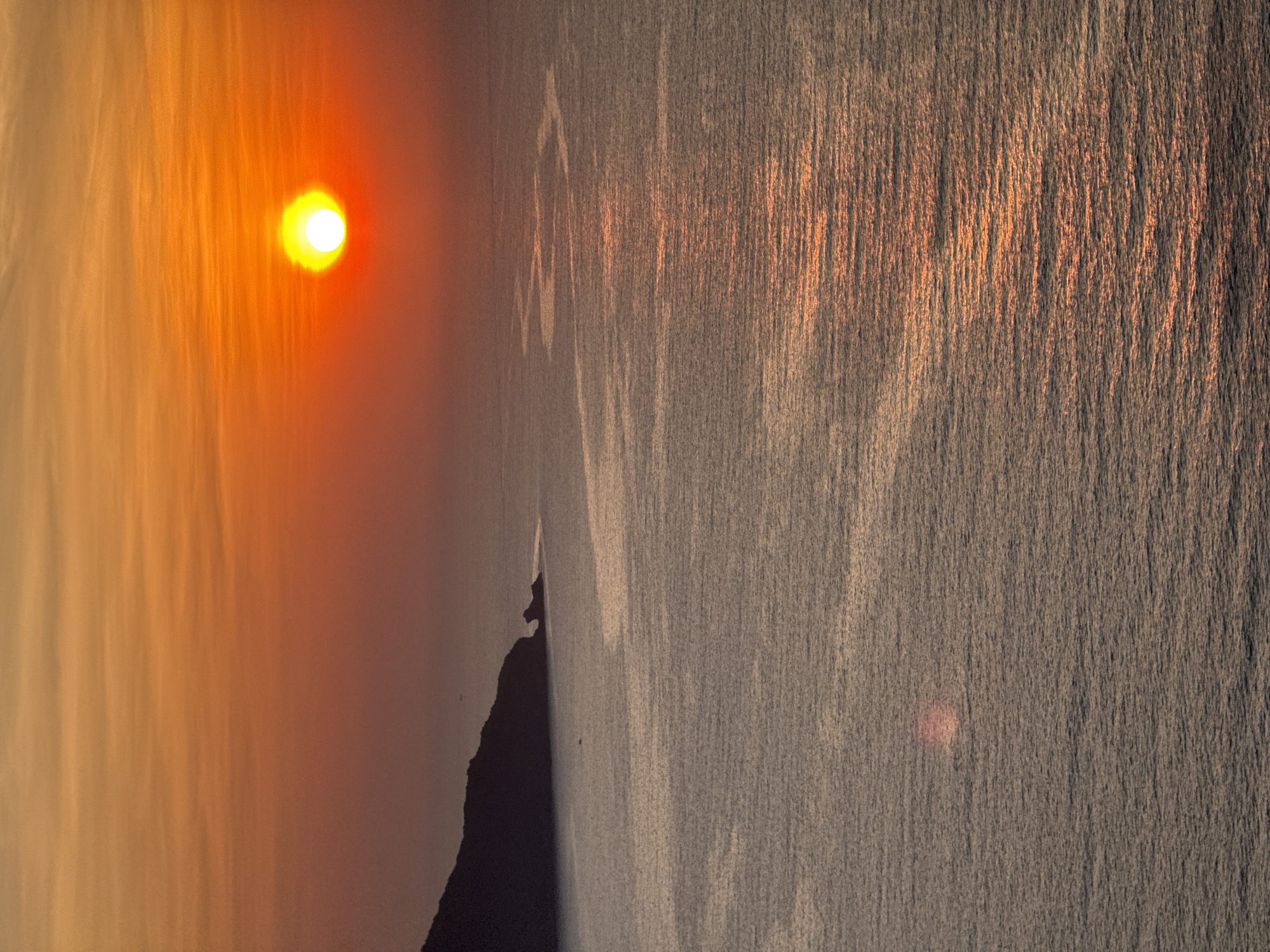 A breathtaking coastal landscape is captured during either sunrise or sunset, showcasing a dramatic interplay of warm light and silhouettes over the waters of Đà Nẵng, Vietnam.
The upper-left portion of the frame is dominated by a radiant, intense sun, glowing with brilliant yellow and orange hues. The sky surrounding it is bathed in a gradient of warm colors, from soft yellows to oranges and faint reds, with hazy, elongated cloud formations stretching horizontally, particularly visible near the sun.
Stretching across the mid-section of the image, a dark, rugged landmass or coastline forms a prominent silhouette. Its irregular, jagged profile suggests cliffs or rocky formations, casting a deep shadow against the bright sky and reflective water. No specific details of vegetation or structures are discernible on the land, emphasizing its stark outline.
The lower half of the image is filled by the sea, which intensely reflects the sun's low light. The water's surface shimmers with countless vertical streaks of orange and yellow light, indicating gentle ripples or small waves catching the sun's rays. This reflection creates a vibrant, textured appearance on the water. A subtle, faint reddish glow or lens flare is visible within the water towards the bottom-right.
The warm, saturated colors and the low position of the sun strongly suggest the scene is either a sunrise or sunset, creating a serene and awe-inspiring atmosphere. There are no visible people, animals, or explicit man-made objects, though a tiny dark speck near the landmass might be a distant bird. No text is visible in the image.