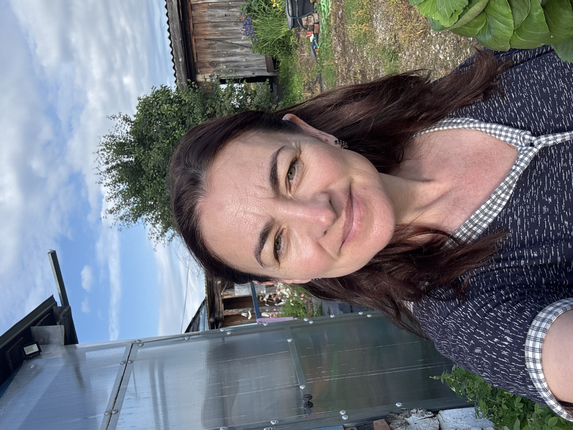 In Urazovo, Russia, a woman with brown hair and fair skin stands outdoors in front of a greenhouse and a wooden shed. She is wearing a dark blue shirt with a checkered collar and cuffs, and she is holding a bunch of green leafy vegetables. The sky is blue with scattered white clouds, and there is some greenery in the background, including a tree and some flowers. The scene appears to be a backyard or garden area.