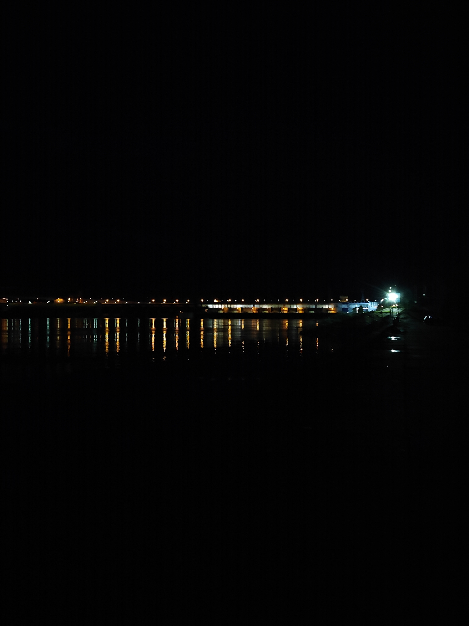 The image displays a nocturnal scene in Chaikovsky, Russia, characterized by a dark body of water reflecting distant artificial lights. The lower two-thirds of the frame is occupied by a dark, calm water surface, producing distinct, elongated vertical reflections of yellow-orange and white light.
Across the midground, a long, horizontal structure, consistent with a dam or a bridge, spans the visible expanse. This structure is illuminated by a series of evenly spaced, warm-toned lights, likely streetlights, forming a bright horizontal line. Below these lights, the darker, solid mass of the structure is partially visible against the night. On the far right side of the midground, a concentrated cluster of bright white lights illuminates a specific section of the structure or an adjacent landmass, possibly indicating a facility or operational area.
The upper third of the image is uniformly black, representing the night sky, devoid of visible celestial bodies or other light sources. No human figures, vehicles, or specific actions are discernible within the frame. The scene presents a static view of illuminated infrastructure over a dark body of water, indicative of a large-scale engineering project such as the Votkinsk Dam on the Kama River, given the location context.
