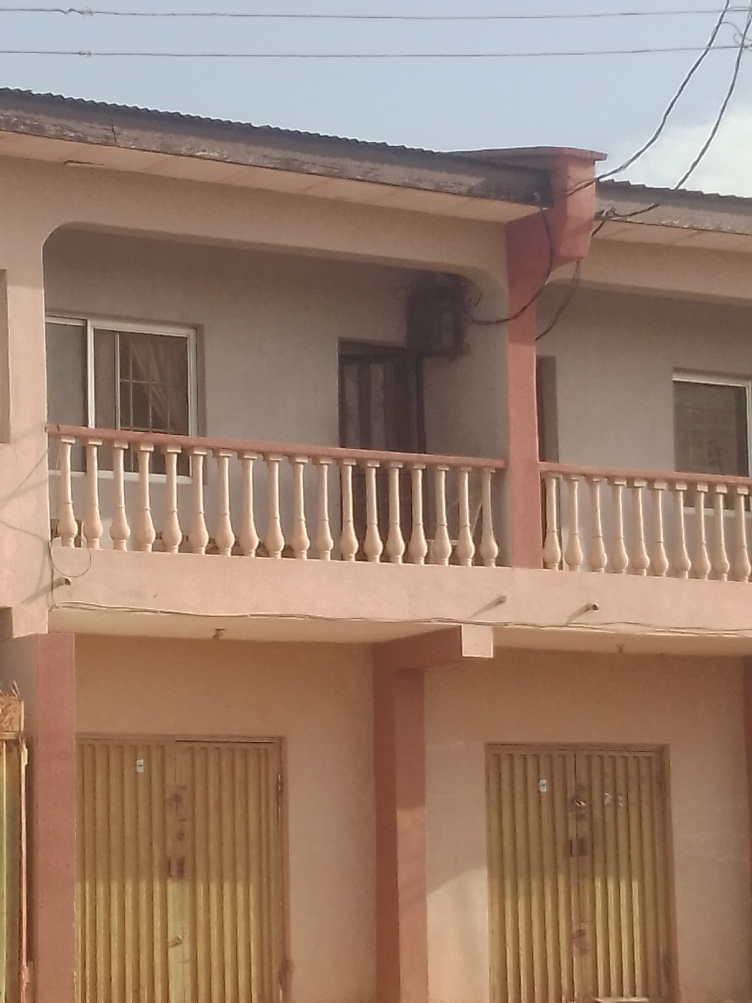 The image shows a two-story building in Sabon Tasha, Nigeria. The building is painted in shades of beige and pink. The upper story has a balcony with decorative balusters. Windows with bars are visible. The ground floor has two closed metal doors. Overhead power lines are present in the sky. An air conditioning unit is attached to the upper story.
