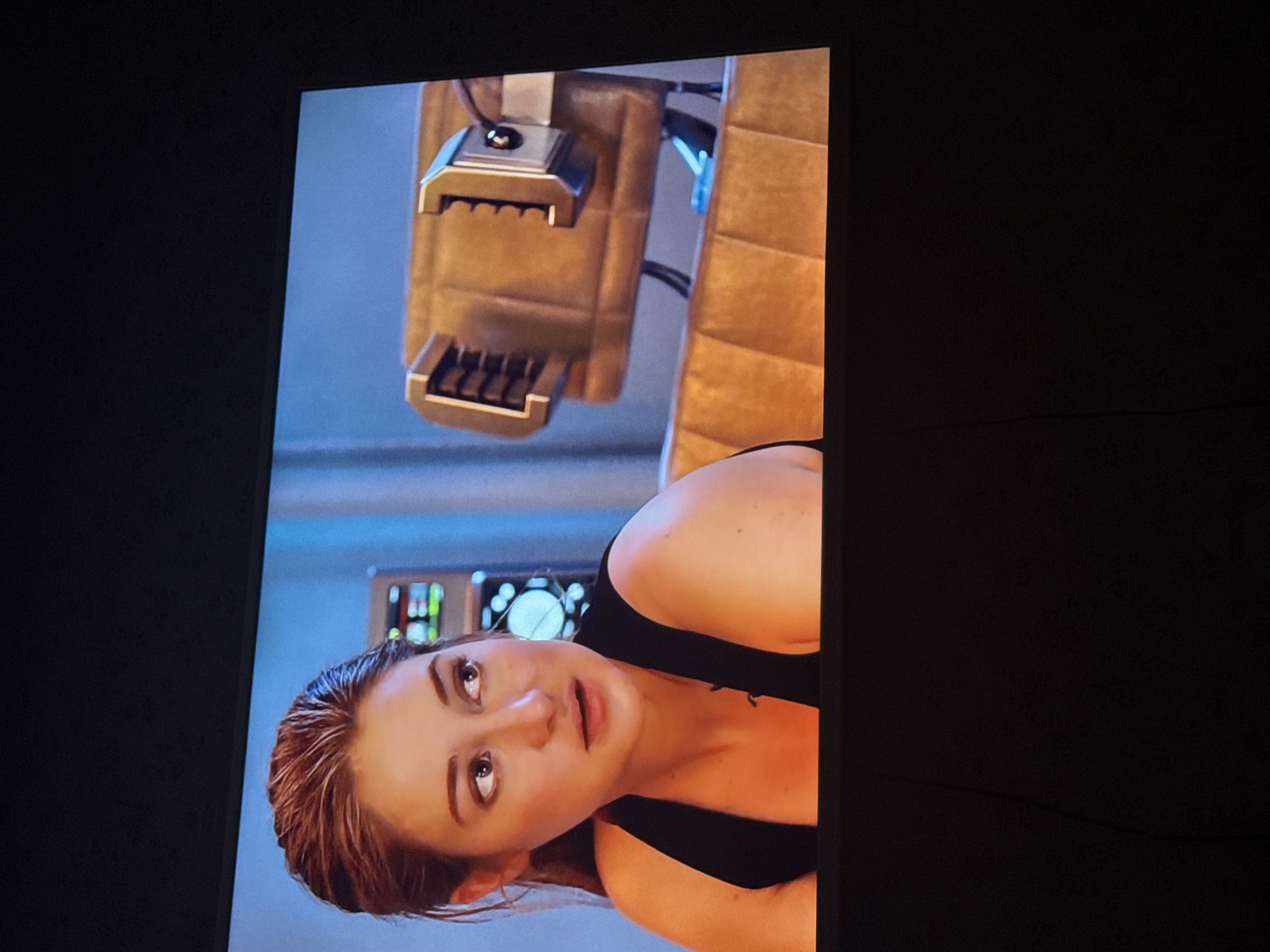 The image shows a woman with light skin and reddish-brown hair looking up with a surprised expression. She is wearing a black tank top. In the background, there is a mechanical device with metallic and tan-colored components. A control panel with various buttons and dials is also visible in the background. The setting appears to be indoors. It is impossible to determine if this image was taken in Mytishchi, Russia, without additional information.