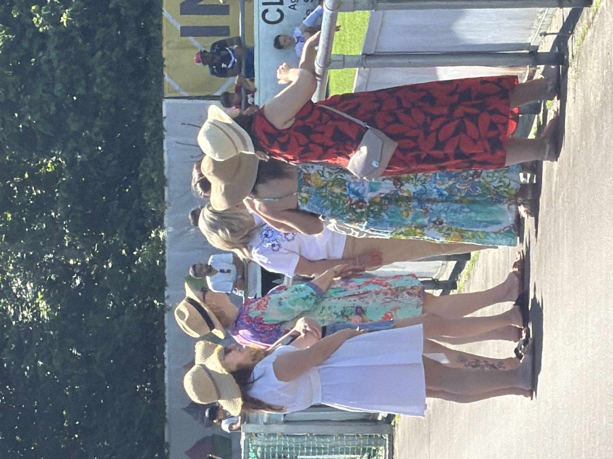 The image depicts a group of five women standing side-by-side, seemingly viewing an event, likely in the city of Fentange, Luxembourg. All are wearing straw hats and dresses. The woman closest to the camera is wearing a white dress, and the next in line is in a light-blue patterned dress. The third woman is wearing a white top and tan pants with a blue-patterned dress, followed by a woman in a blue-floral print dress. The woman furthest from the camera is wearing a red floral-print dress and carrying a small grey purse. They are standing alongside a low metal barrier. People and signage are visible in the background. A dense tree line is on the far left.