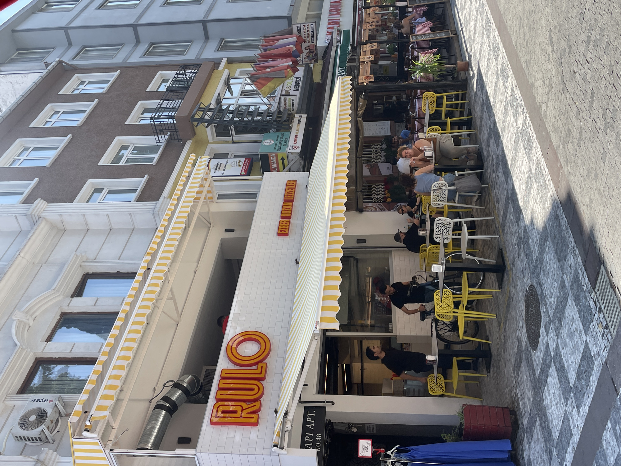 The image shows a street scene in Kadıköy, Turkey, with several restaurants and shops visible. In the foreground, an outdoor seating area of a restaurant with yellow chairs and tables is set up on a cobblestone pavement. People are seated at the tables, some eating and conversing. A man in a black shirt is standing near the entrance of the restaurant. Above the seating area, a restaurant called "RULO" is indicated by a prominent red sign. The building facade features a white tiled exterior and a yellow and white striped awning. Further down the street, other businesses and buildings are visible, including those with Turkish and German signage, suggesting a mix of local and international establishments. The scene is bathed in sunlight, casting shadows on the pavement.