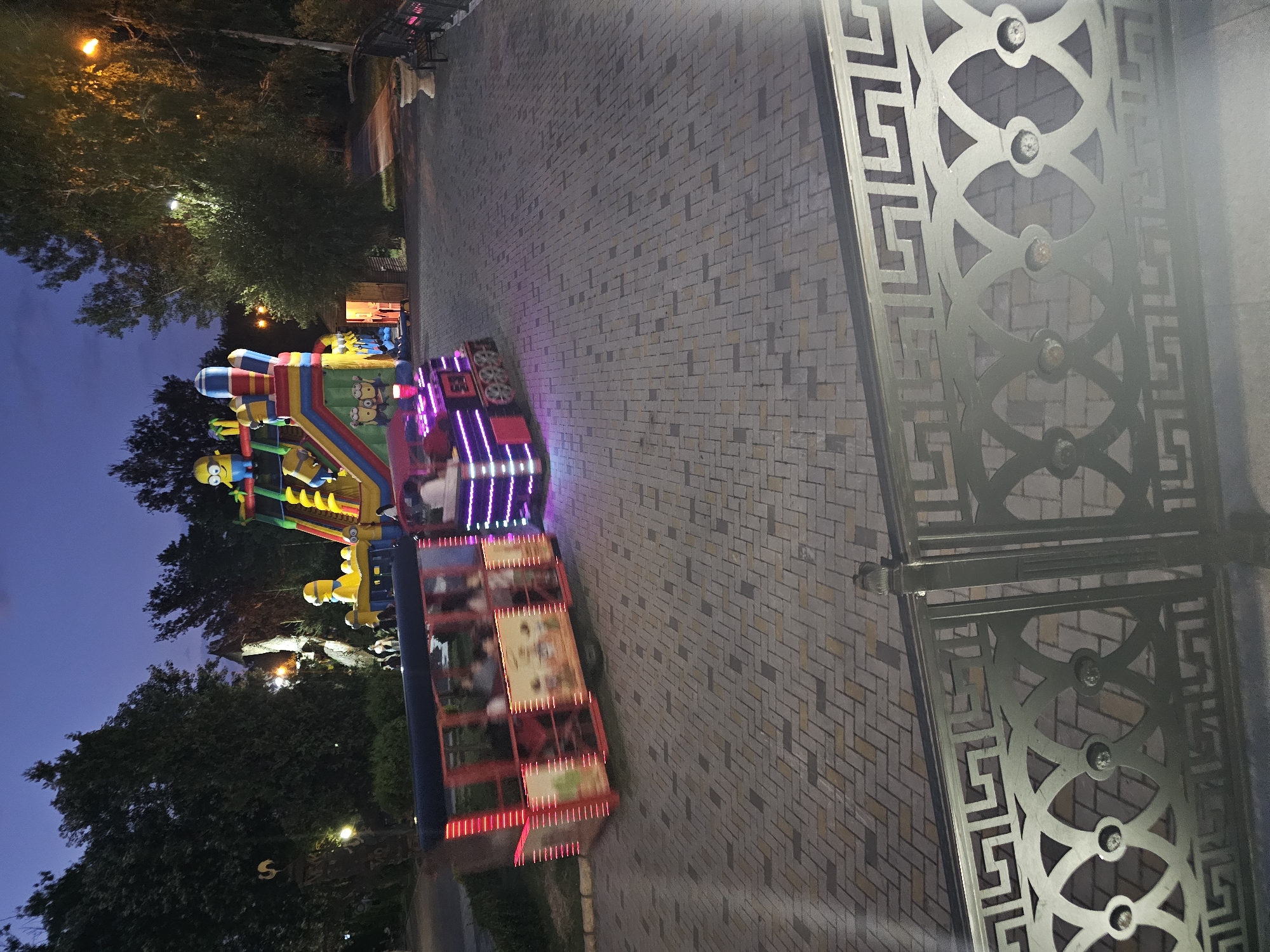 The image depicts an amusement park scene at dusk in Cherkessk, Russia. A small, red amusement train with illuminated purple lights on its engine and red lights along its carriages is moving on a paved path. The train appears to have passengers inside. In the background, a large inflatable structure with characters resembling Minions is visible, along with trees and streetlights. A decorative metal railing is in the foreground. The sky is a deep twilight blue.