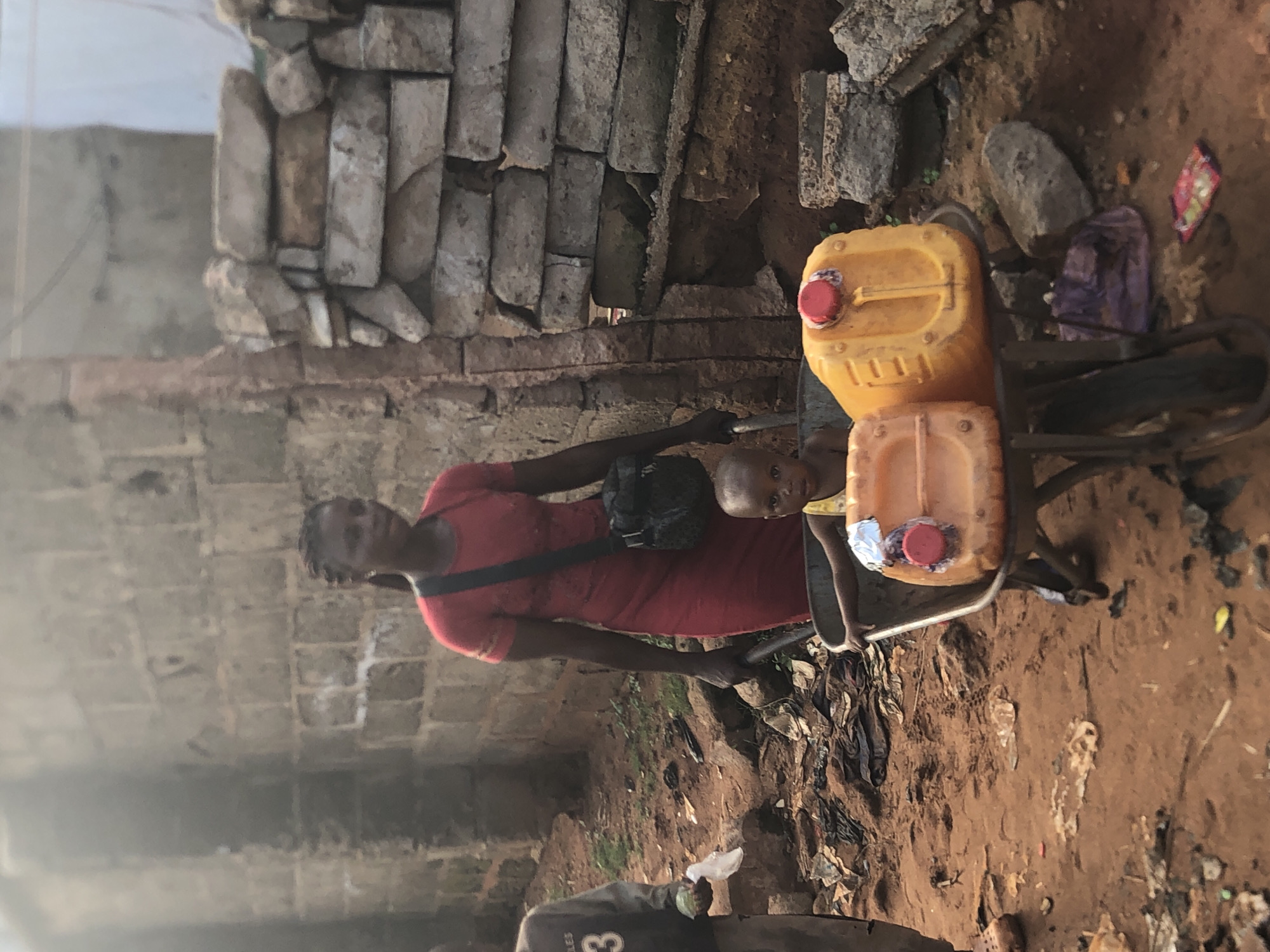 The image, taken in Ukwu-Abwa, Nigeria, captures a woman and a child with a cart carrying water containers. The scene is set outdoors in what appears to be a residential or communal area, characterized by a narrow passage between rough-hewn stone and brick walls.
The primary subjects are an adult woman and a young child. The woman, wearing a red dress and a black crossbody bag, stands on the left side of the frame, holding the handle of a metal cart. Her gaze is directed slightly to her left. A young child, possibly a toddler, stands next to the woman and the cart, wearing a light-colored top, and looks towards the viewer.
The central object is a metal two-wheeled cart designed for transport, possibly a hand truck or trolley. It carries two yellow plastic jerrycans, likely filled with water or other liquids, both secured on the cart. The larger jerrycan is positioned above a smaller one, and both have red caps.
The setting features an uneven, reddish-brown dirt ground, scattered with small rocks, patches of sparse vegetation, and some litter, including plastic bags. The background consists of robust but weathered walls constructed from various sizes of irregular stones and bricks, indicating a simple or older building style.
The activity depicted is consistent with daily life in the region, likely involving the transport of water. The lighting suggests it is daytime, with no harsh shadows indicating strong direct sunlight. The expressions on the faces of the woman and child appear neutral. No significant text is clearly legible, though a faint "3" and other indistinct markings are visible on a dark object partially cropped in the bottom foreground.