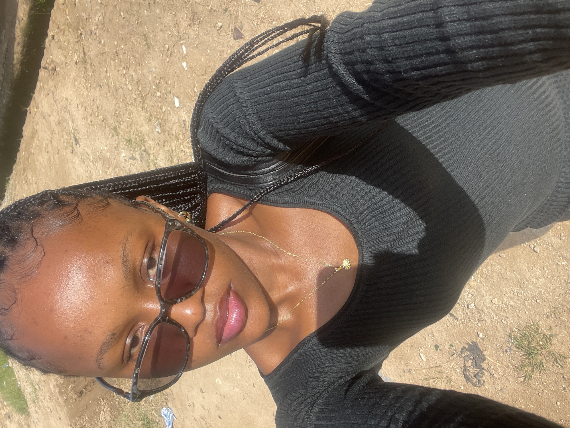 A young Black woman with braided hair, wearing sunglasses and a black ribbed top with a gold necklace, poses for a selfie outdoors. The background appears to be a dusty, unpaved area with sparse vegetation, consistent with the arid environment of Jos, Nigeria. The lighting suggests it is daytime.