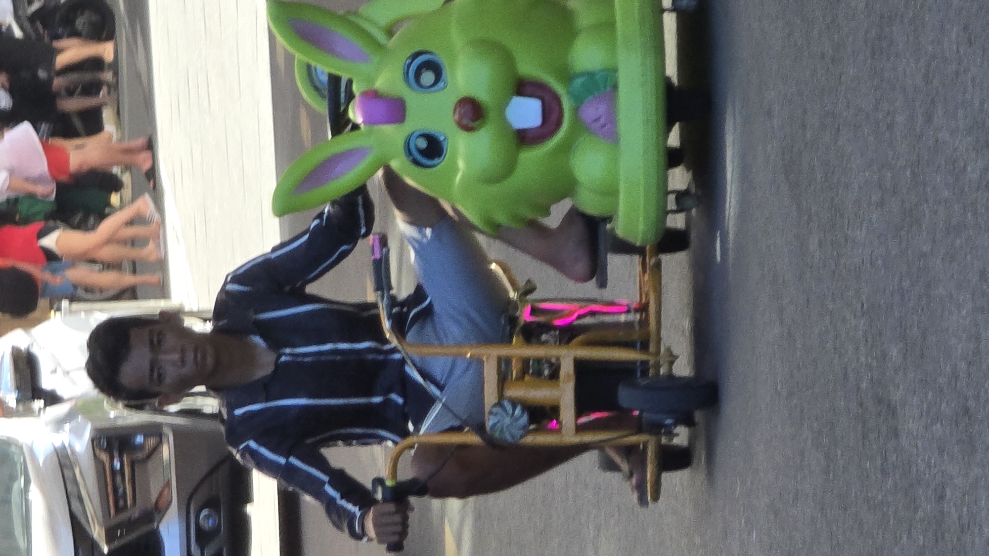 A person is riding a low-lying, homemade, yellow-framed vehicle with a large, green rabbit head affixed to the front. The person is wearing a blue and white striped shirt and light gray shorts. The vehicle has a small, black front wheel and appears to have pink lights underneath. The person is gripping black handlebars. The background includes a blurred sidewalk with several people and a portion of a white vehicle, possibly a car or truck. The location is identified as Bac Nha Trang Ward, Vietnam. The surface under the vehicle appears to be asphalt.