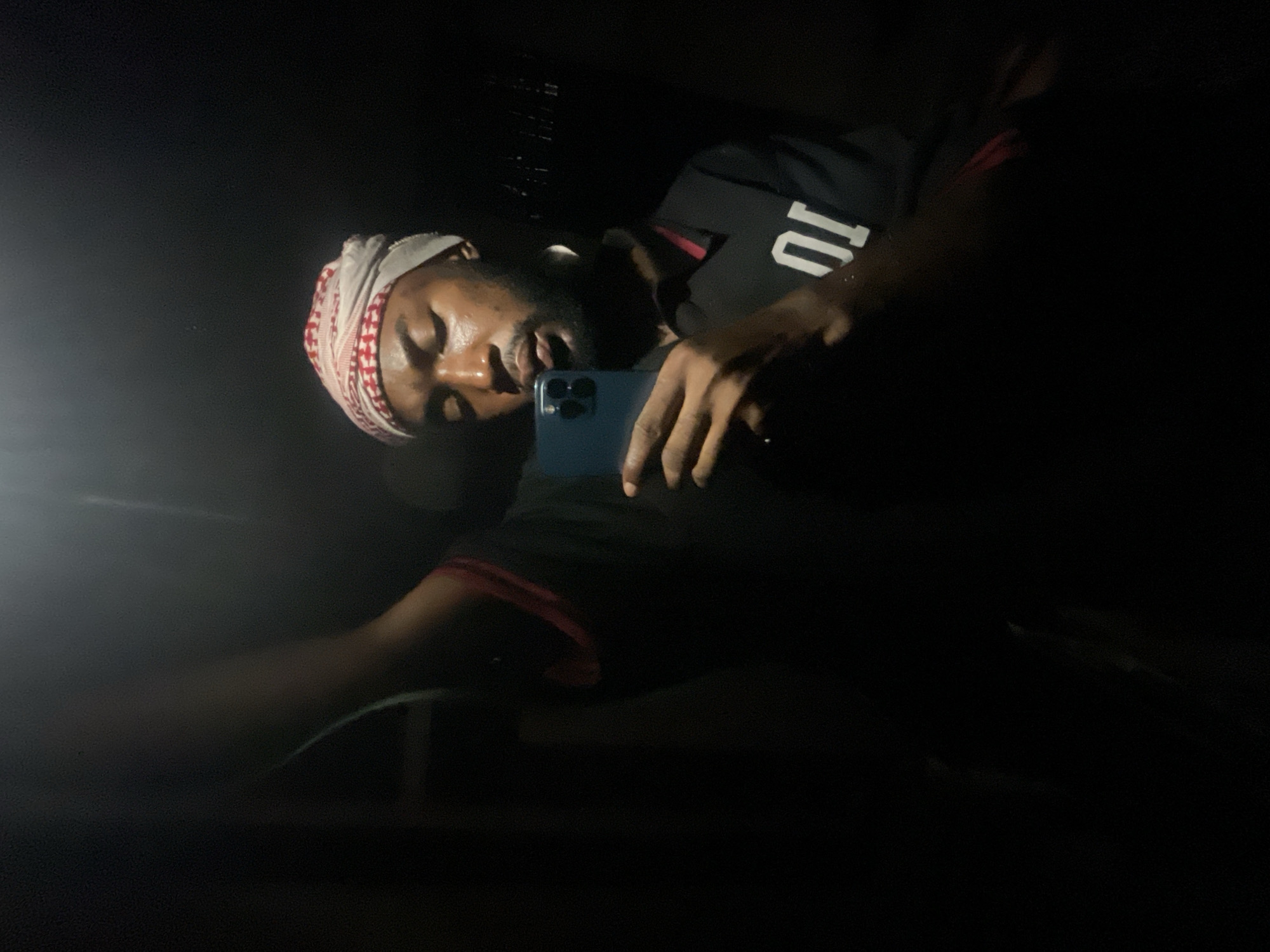 A person, presumably male, is illuminated by a light source to the left of the frame. He is wearing a patterned head covering and a dark top with a white number "10" visible. The person is holding a blue smartphone. The environment appears to be dark, possibly indoors. There is no information to verify or deny if the image was taken in Lokoja, Nigeria.