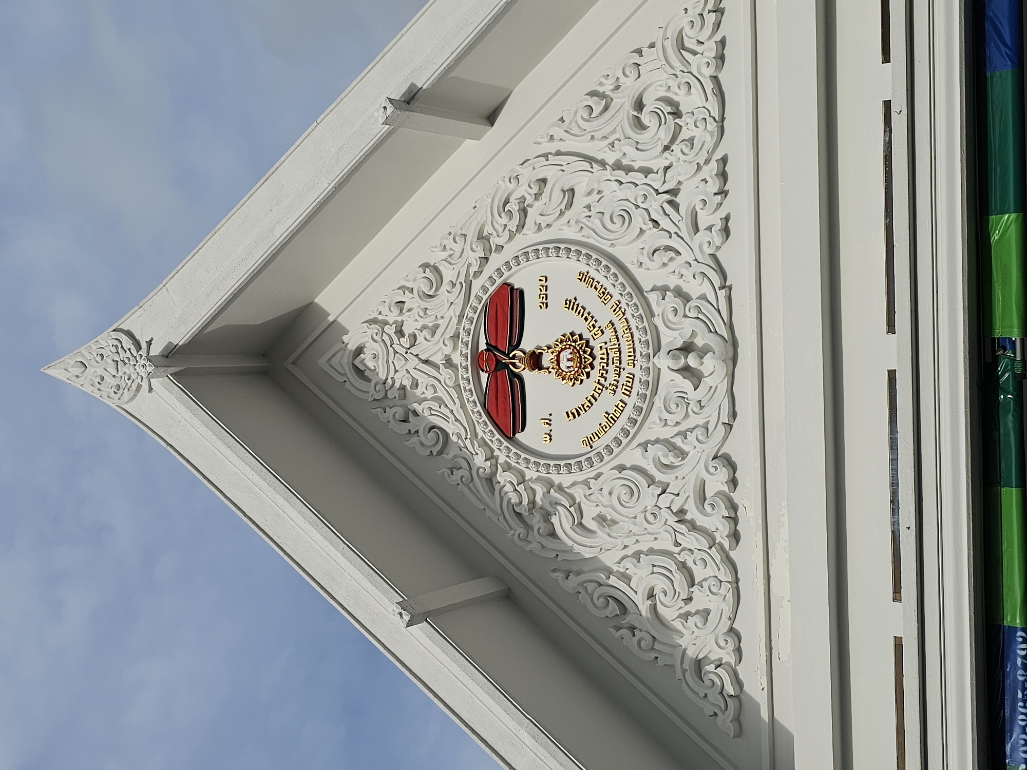 The image displays the upper facade of a white building under a blue sky with scattered clouds. The building features intricate white decorative carvings in a pediment, centered around a circular emblem. The emblem contains a gold medallion with Thai inscriptions and a red ribbon. To the right of the emblem, the Thai numeral "2520" is visible in gold. Below the decorative facade, there are horizontal white bands and a hint of colorful material, possibly banners or signage, at the very bottom of the frame, with a partial view of a phone number "02-865-8792" visible. The architectural style suggests a Thai temple or government building. Without external context or visible landmarks, the specific location within Bangkok, Thailand, cannot be determined.