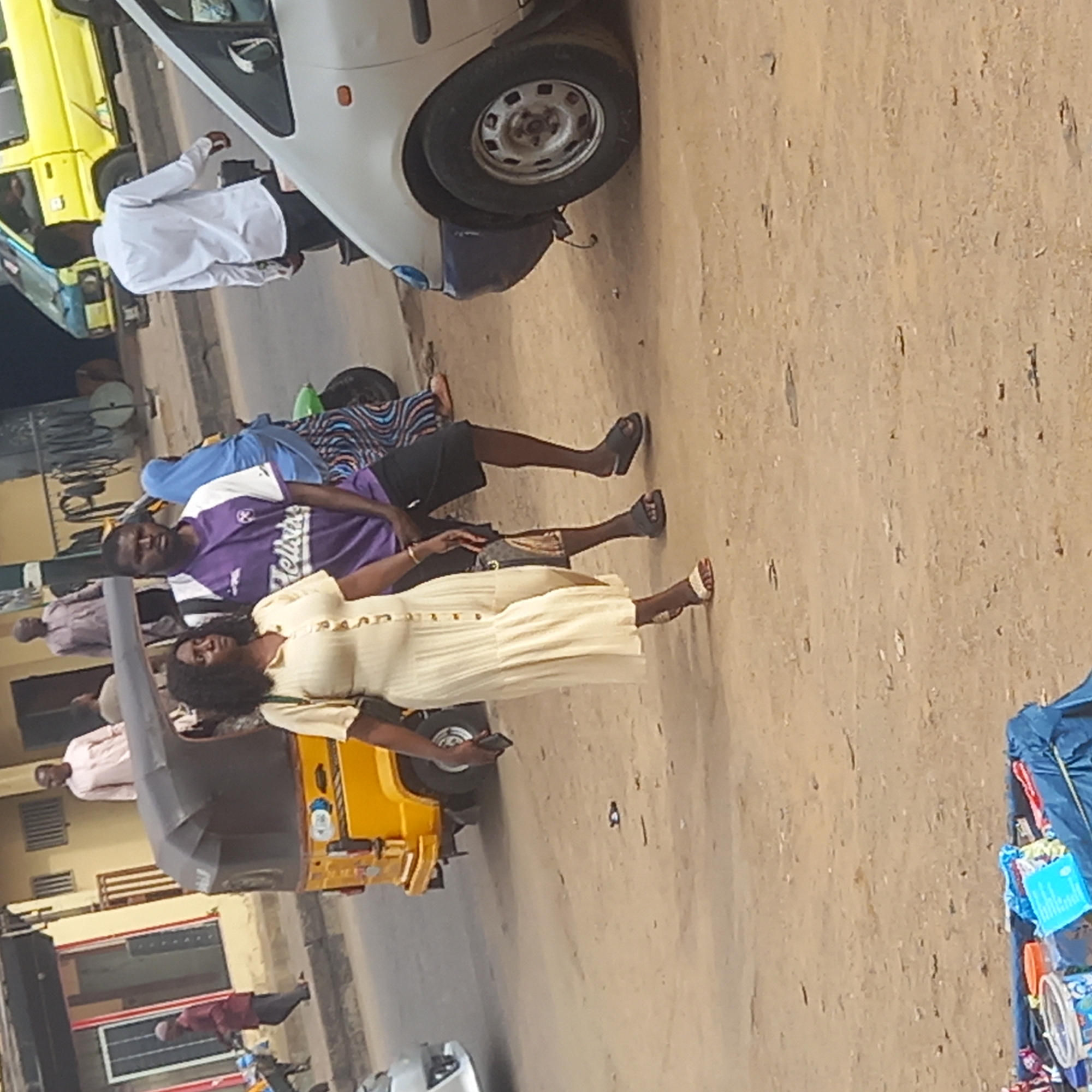This image, taken in Sardauna, Nigeria, depicts a daytime street scene with several individuals and vehicles. The ground is a light brown, dusty surface, characteristic of unpaved or heavily trafficked roads in the region.
In the center of the frame, two people are standing. A man, wearing a purple and white t-shirt with "Hellsun" (or similar text) visible on it, and dark shorts, stands with dark-colored flip-flops on his feet. He looks directly towards the camera. To his right and slightly forward, a woman in a long, light yellow or cream-colored pleated dress with buttons is holding a dark smartphone in her hand. She has dark, curly hair and wears light-colored sandals. Her gaze is directed slightly away from the camera.
Various vehicles are present, contributing to the busy street atmosphere. A white car, likely a sedan, is parked or stopped on the left, with a person dressed in a white tunic standing nearby. Further to the left, a portion of a yellow and blue commercial vehicle is visible. In the lower left, part of a yellow tricycle, commonly known as a "Keke Napep" or "Keke Marwa" in Nigeria, is seen. In the background, other people are faintly visible, including one in a light pink shirt and another in a maroon shirt, suggesting a populated area. On the far right, a blue tarp or bag appears to hold an assortment of small items, possibly goods for sale. The overall scene conveys a snapshot of daily life in a Nigerian town or city.
